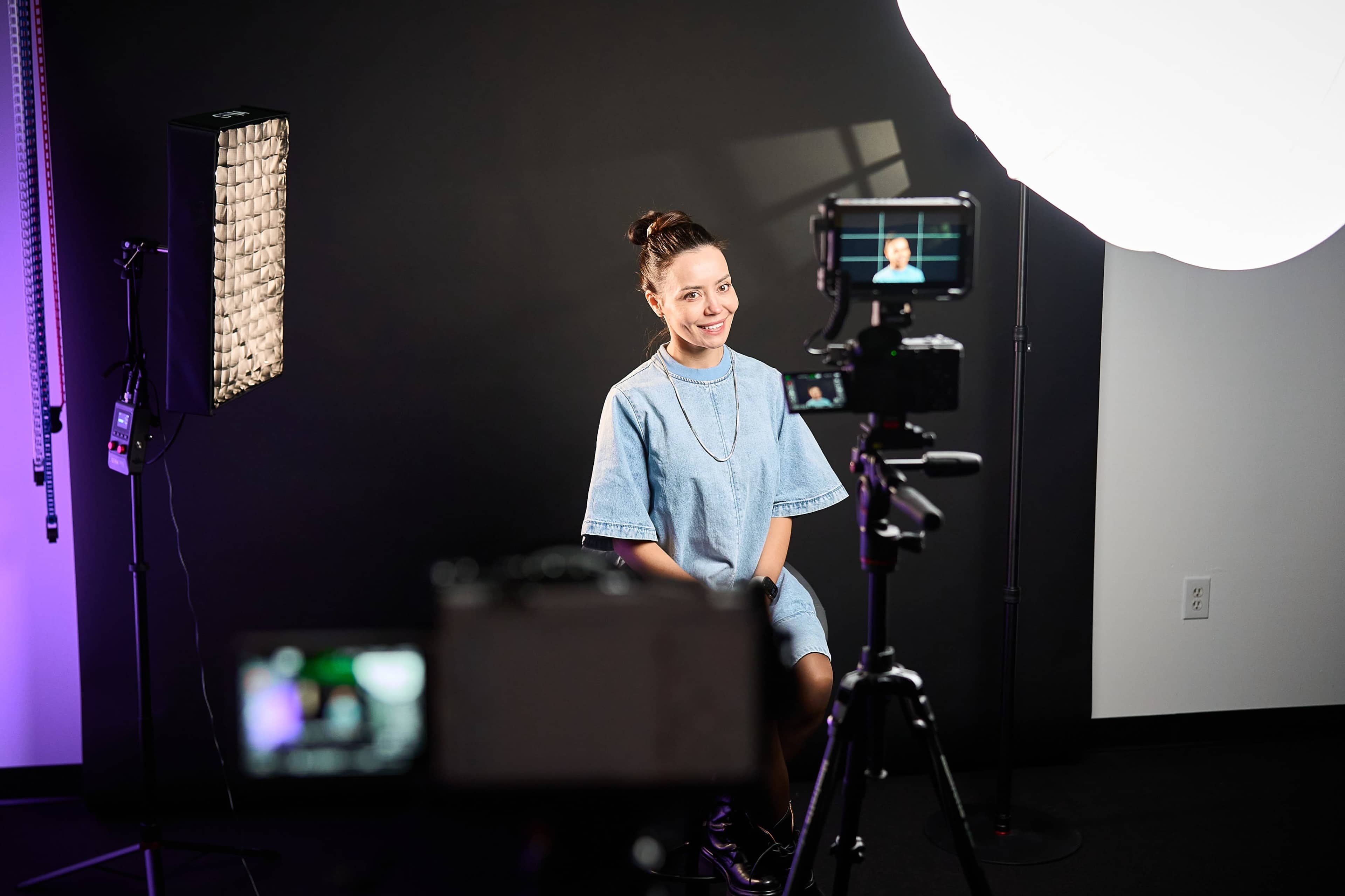 A woman in a denim dress sits in front of cameras and lighting equipment in a studio setting.