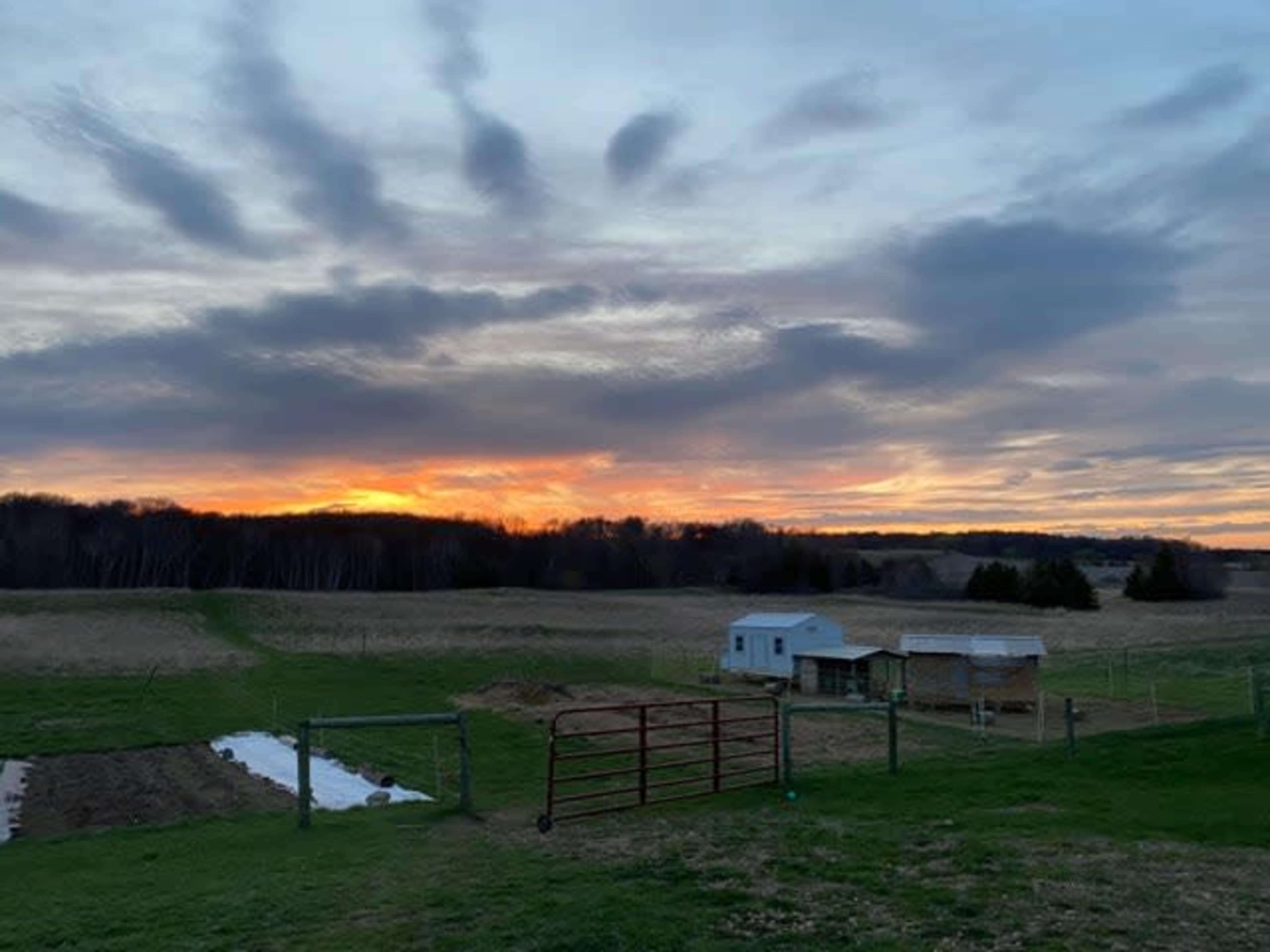 The image shows a rural landscape at sunset, featuring a small farmhouse, a barn, and fields under a cloudy sky.