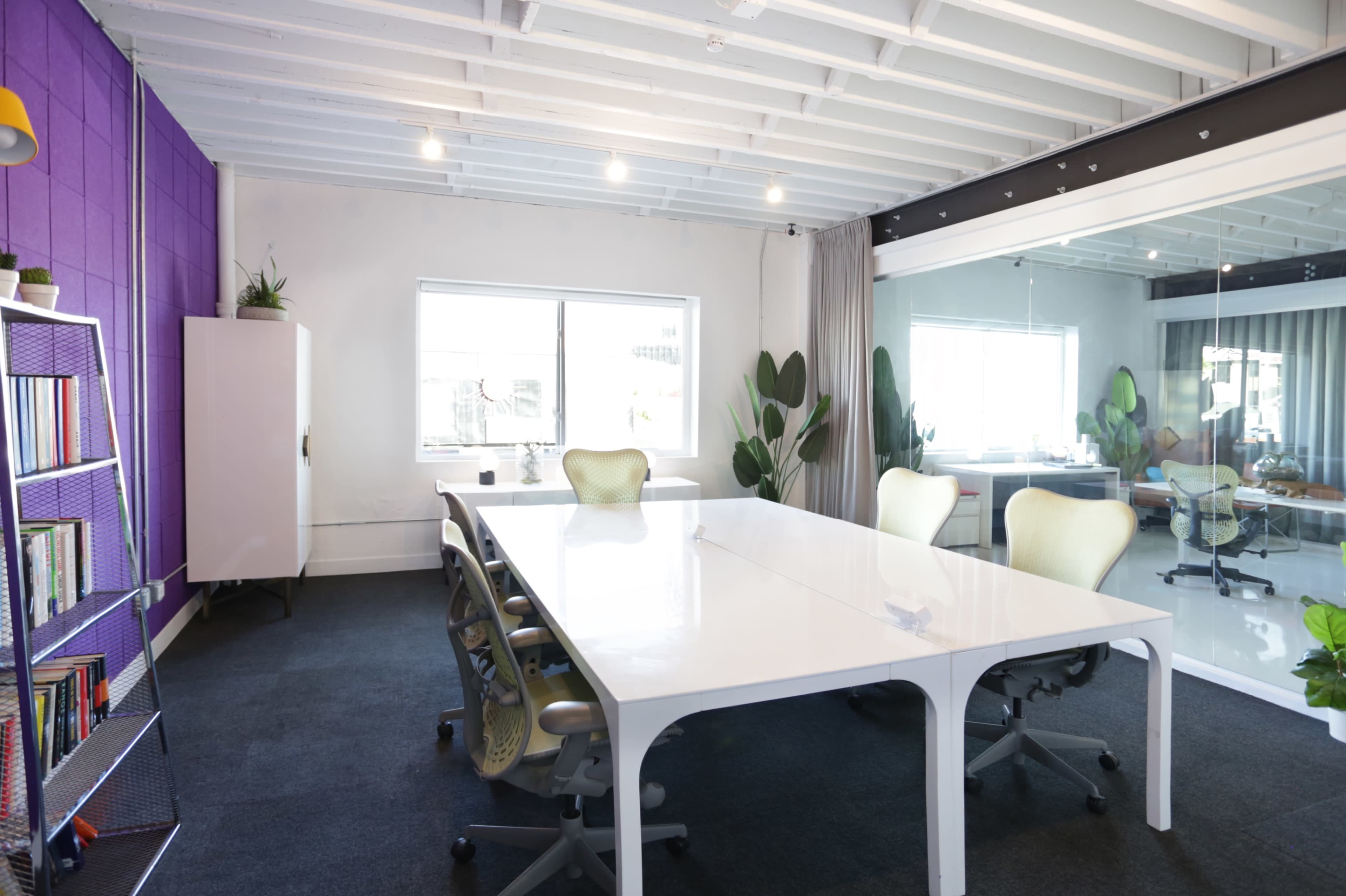 A modern office space features a large white conference table surrounded by ergonomic chairs, with purple accent walls and plants positioned throughout the room.