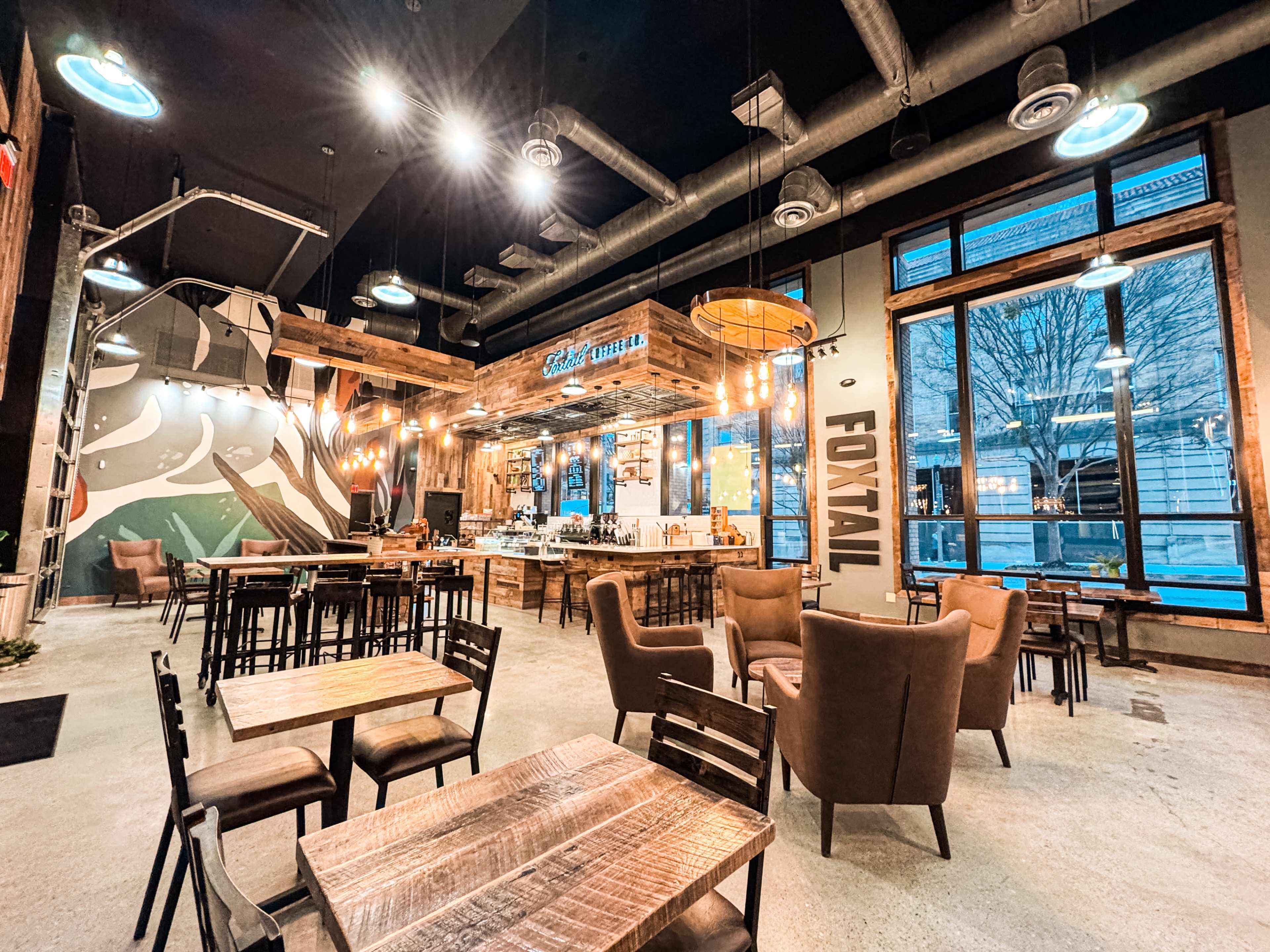 The image depicts a modern café interior with wooden furniture, artistic wall murals, and large windows allowing natural light to enter.