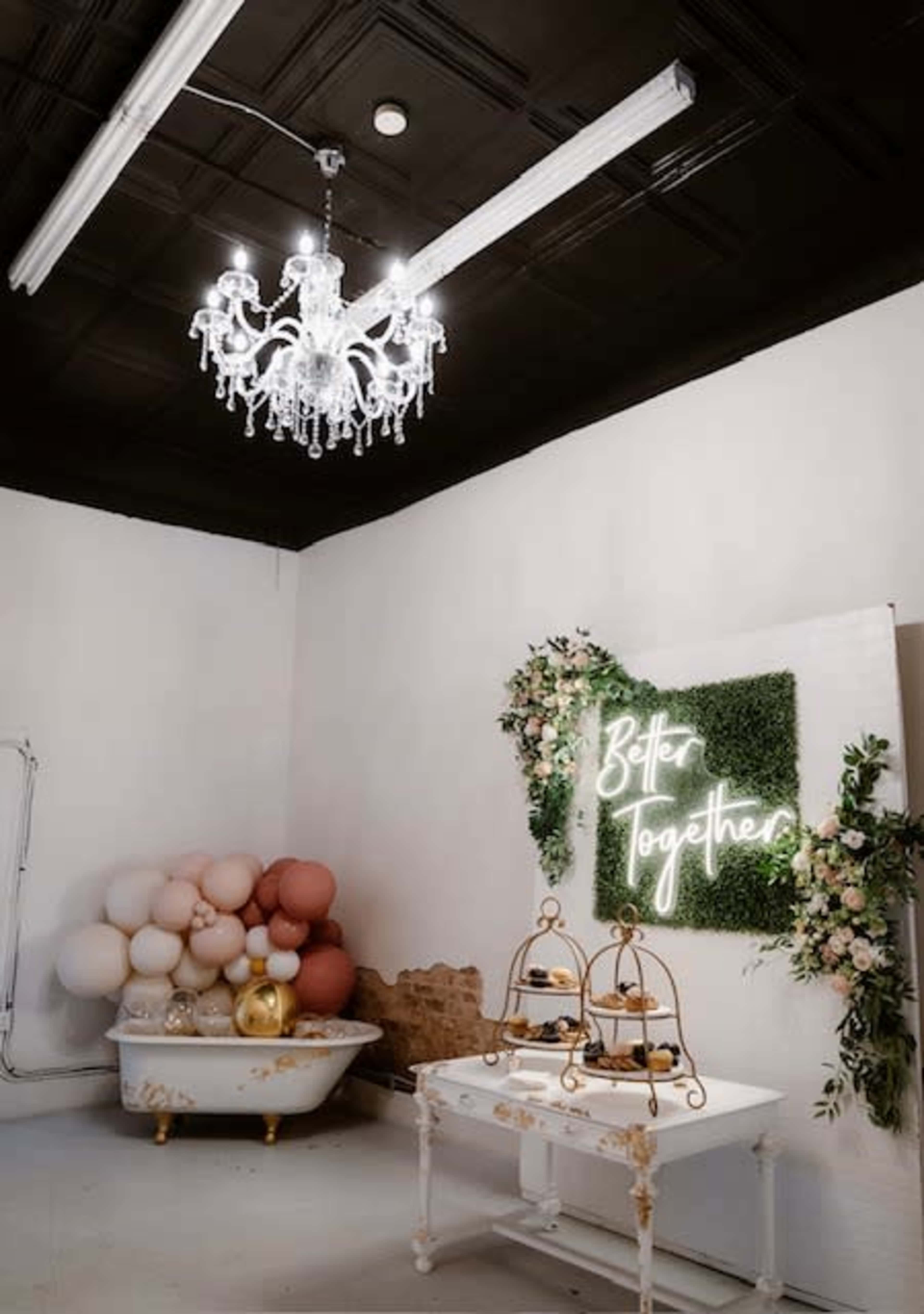 The image shows an elegant indoor space featuring a vintage bathtub, a decorative dessert display on a white table, and a neon sign that reads "Better Together," surrounded by floral accents and a backdrop of greenery.