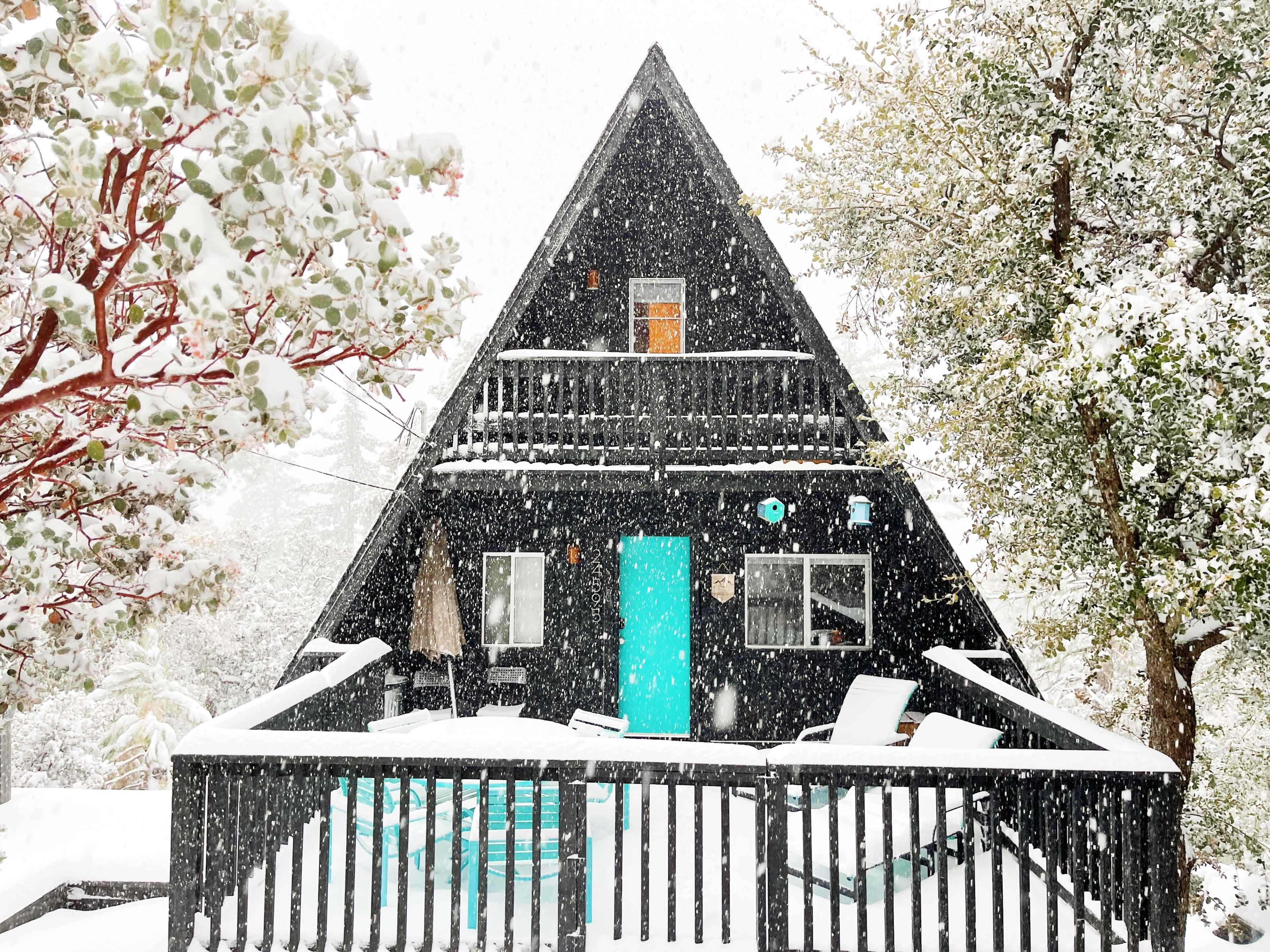A black A-frame cabin with a turquoise door is surrounded by snow-covered trees in a winter landscape.