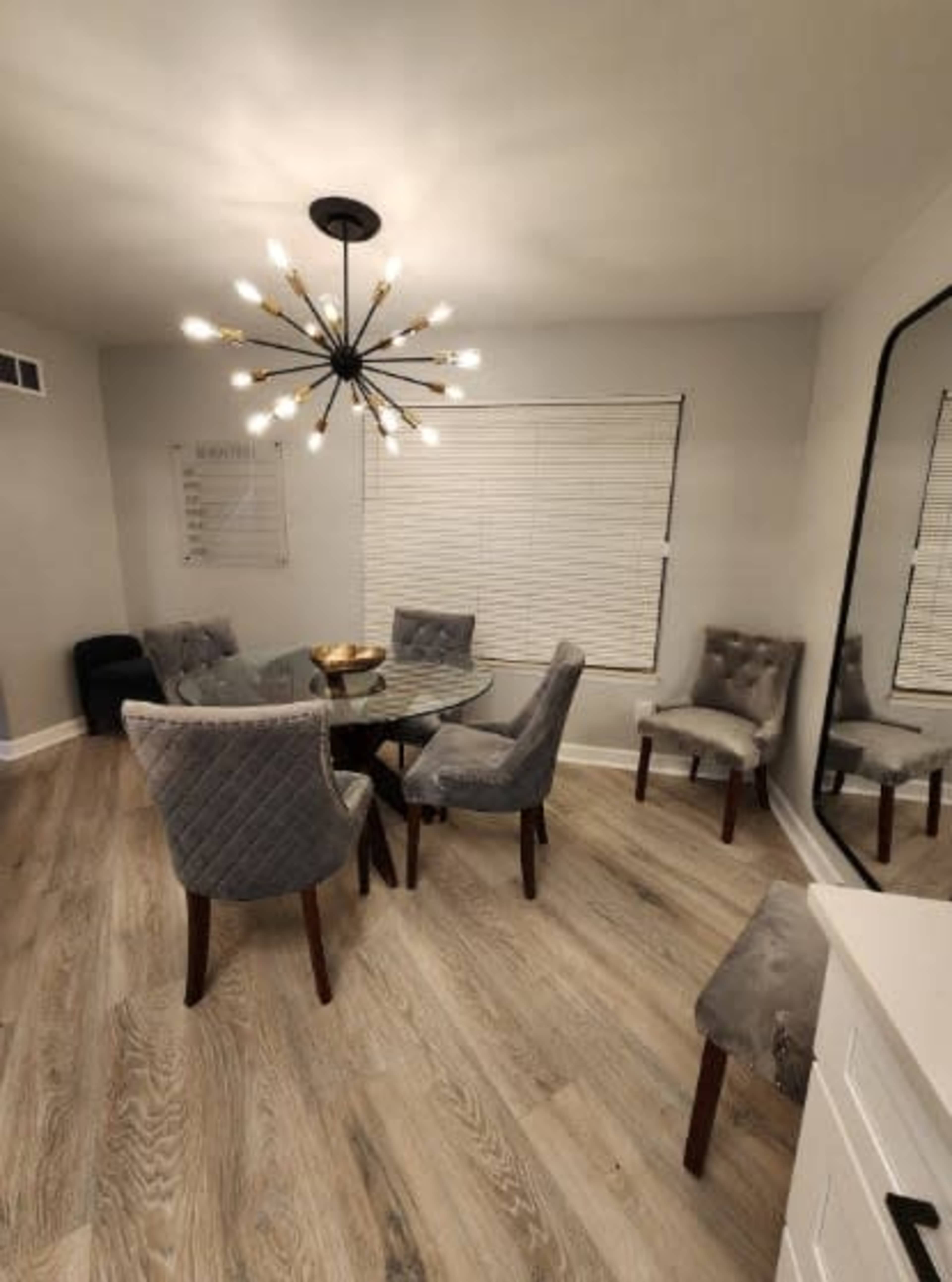 A modern dining area featuring a glass table surrounded by six upholstered chairs, with a large chandelier overhead and a mirrored wall.