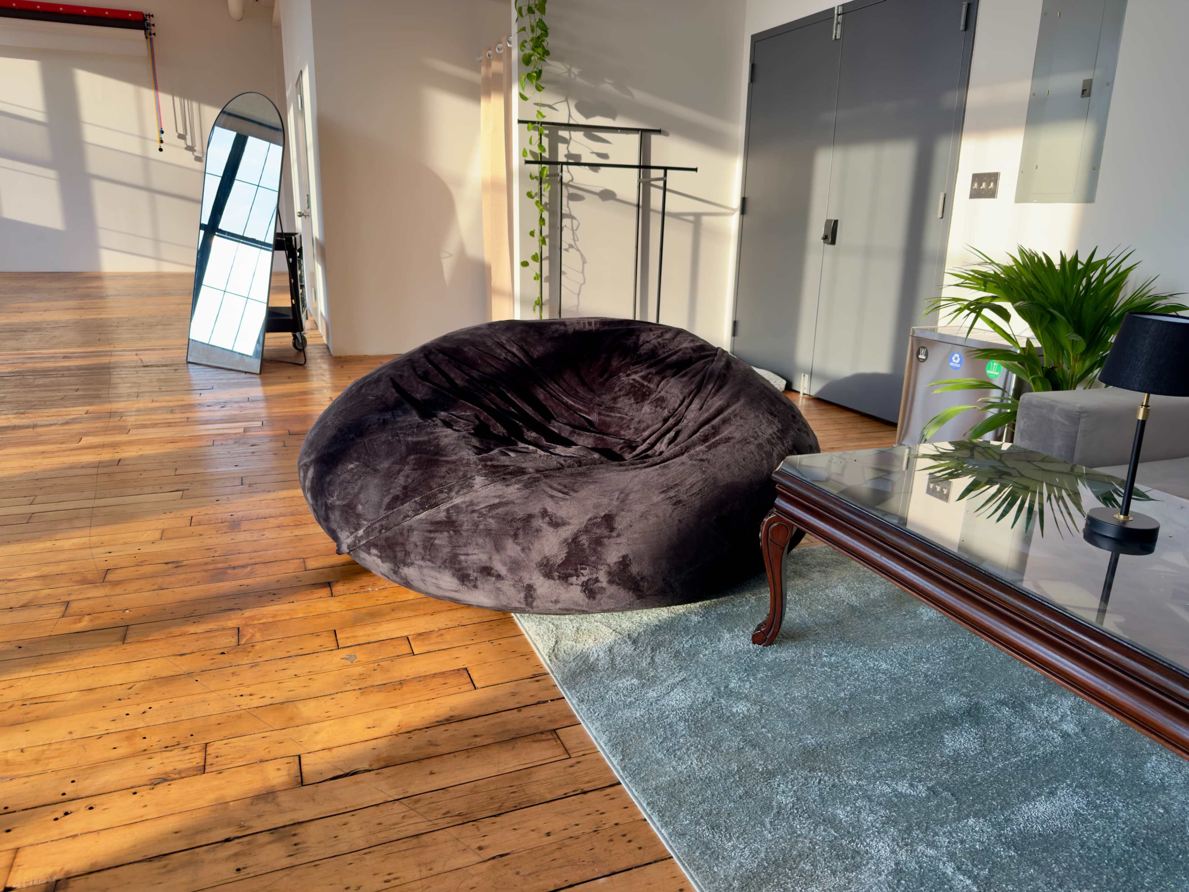 The image shows a spacious room with wooden floors, featuring a large black bean bag chair, a glass coffee table, and plants along the walls.