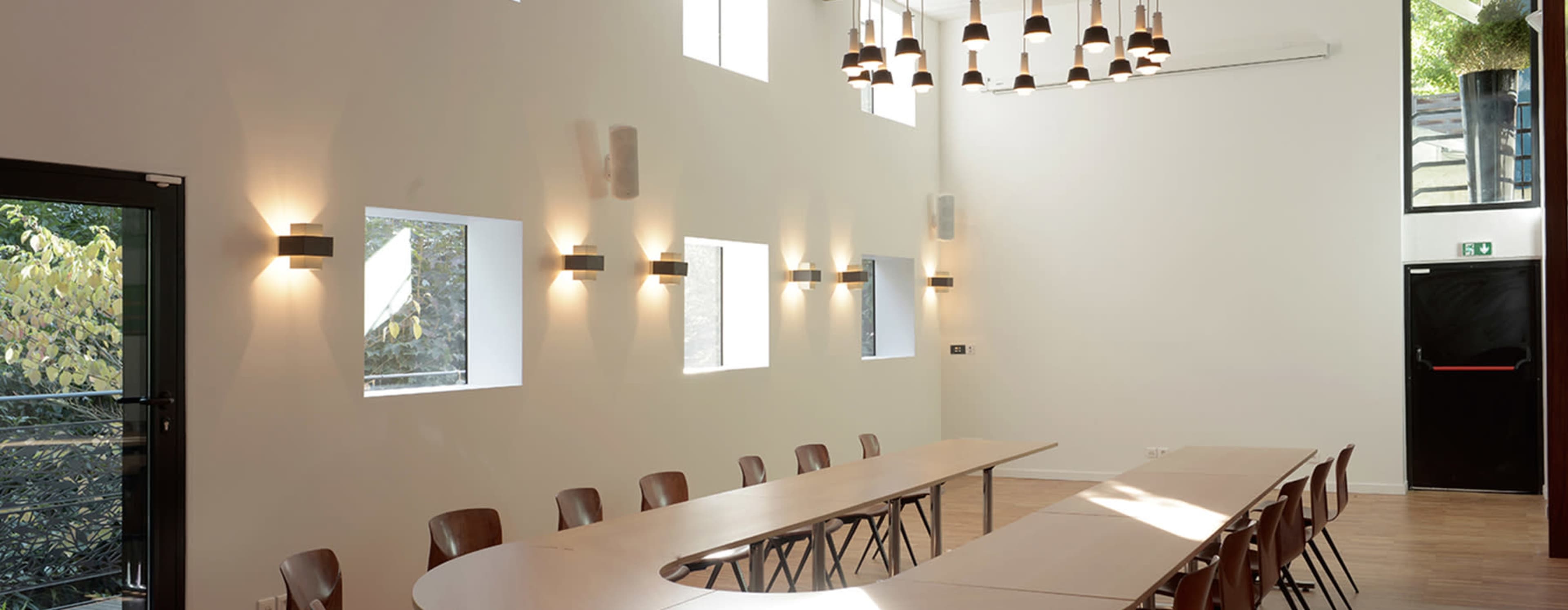 A bright meeting room with a long, curved table, multiple wall-mounted lights, and large windows allowing natural light to enter.