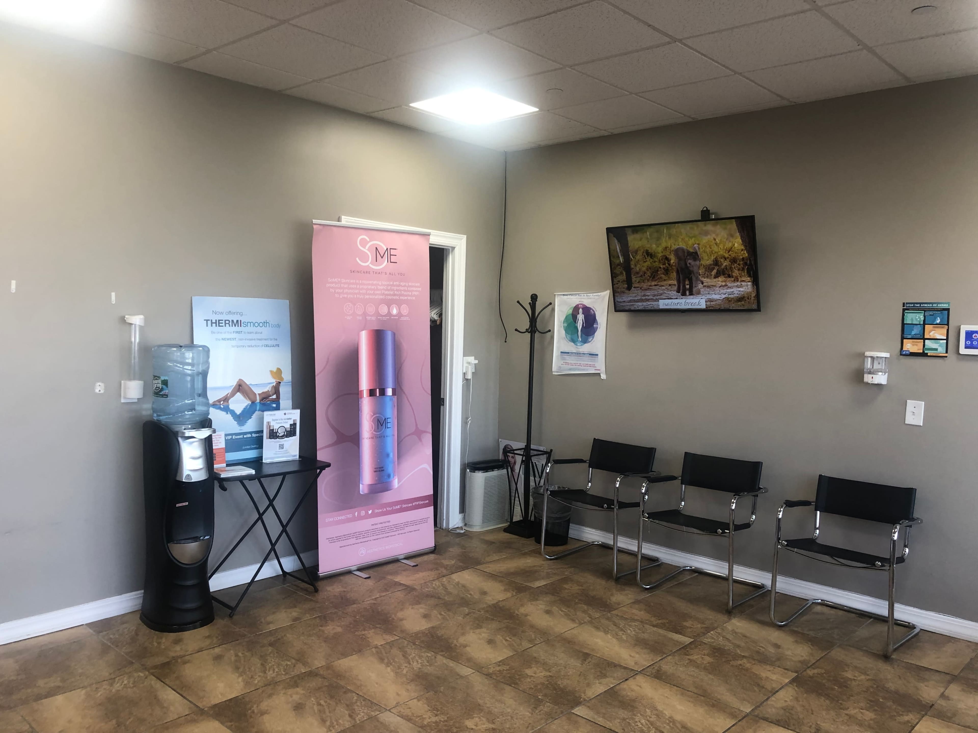 The image shows a waiting room with a water cooler, a banner for a skincare product, a television displaying a nature scene, and several black chairs arranged for seating.