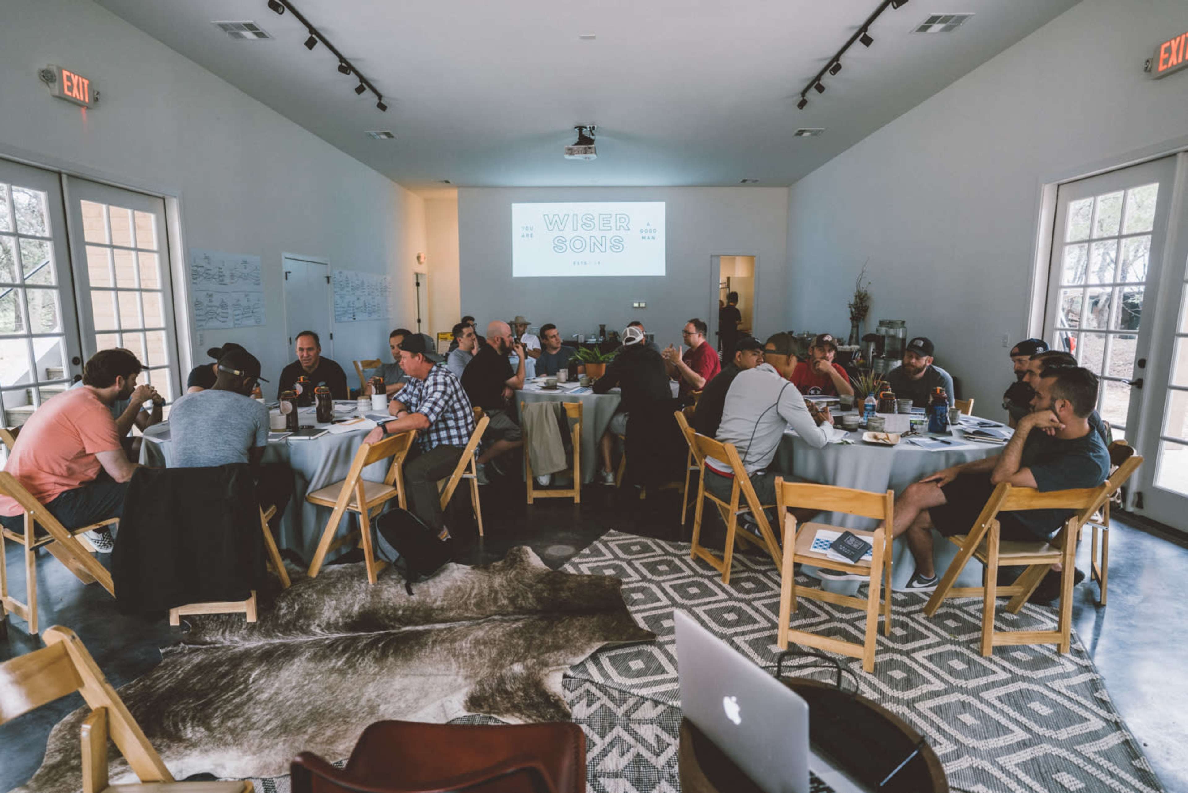 A group of approximately 30 people is seated around several tables in a well-lit meeting room, engaged in discussion with materials and laptops in front of them.