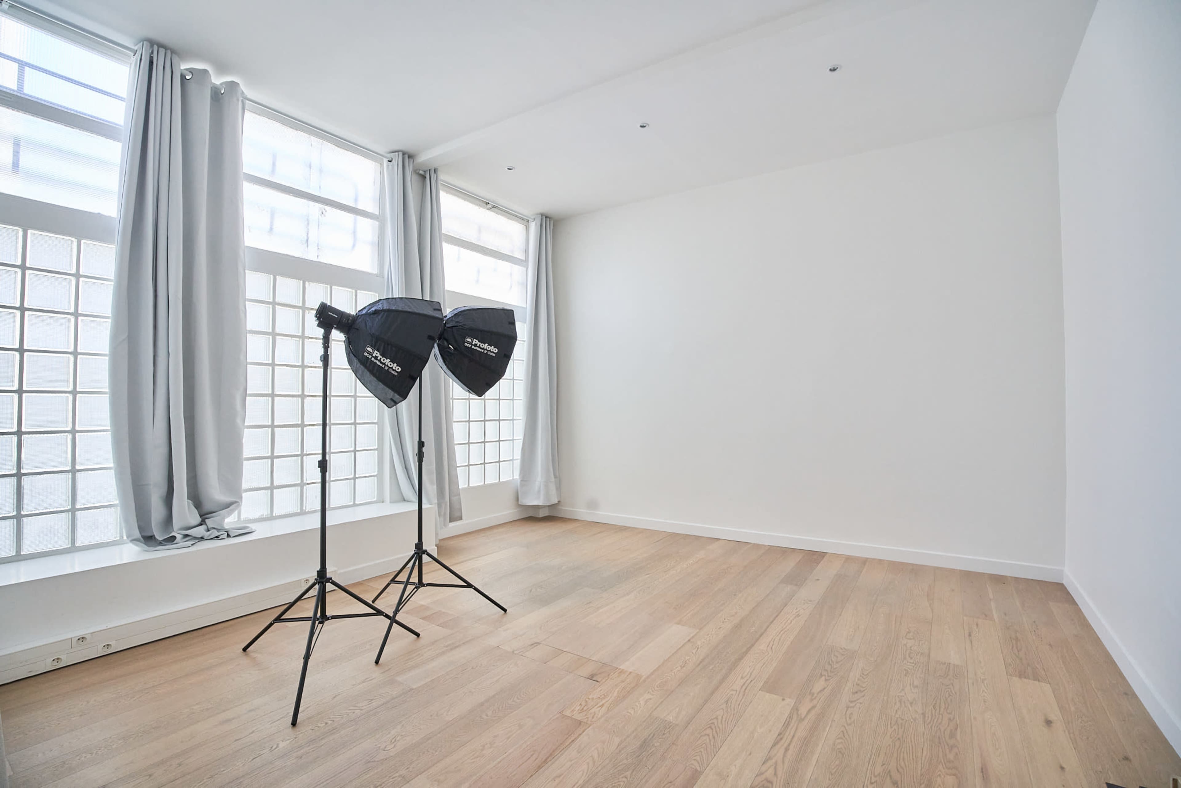 A bright, empty room with large windows and two lighting softboxes on stands.