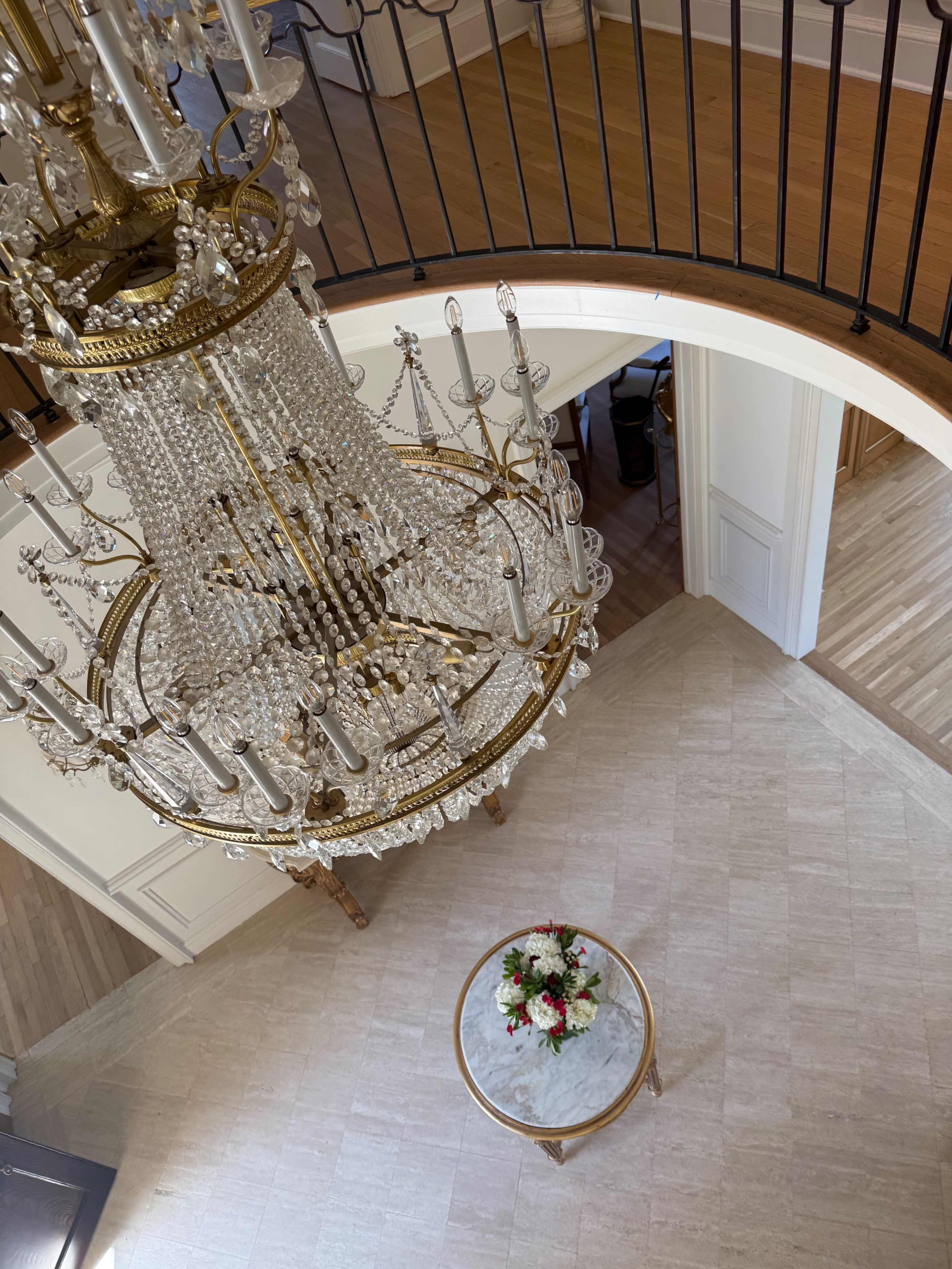 A large crystal chandelier hangs above a round table adorned with a floral arrangement in a spacious foyer.