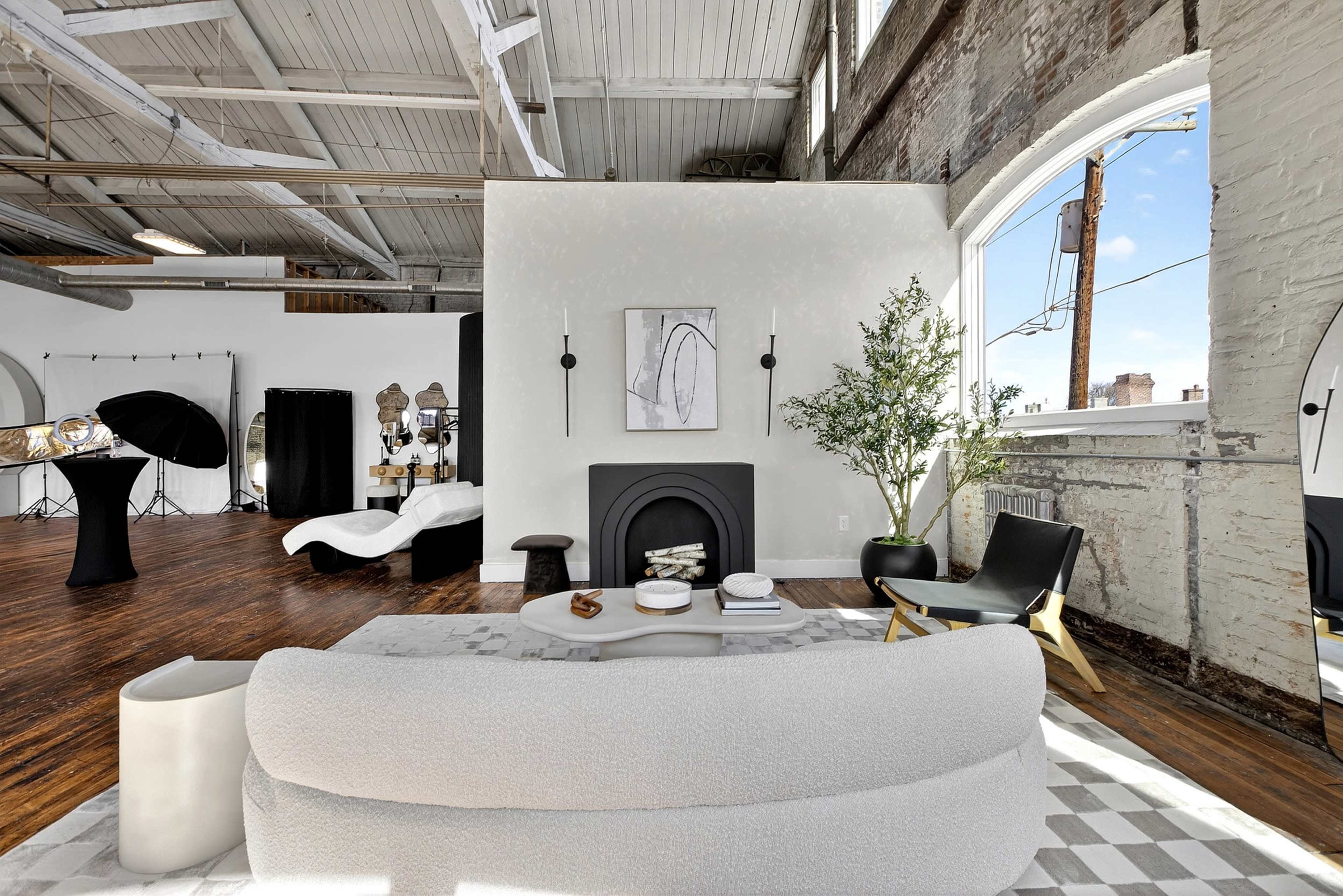 The image shows a spacious, modern loft with a light-colored sofa, a fireplace, and a large window that allows natural light to enter, while a photography setup is visible in the background.