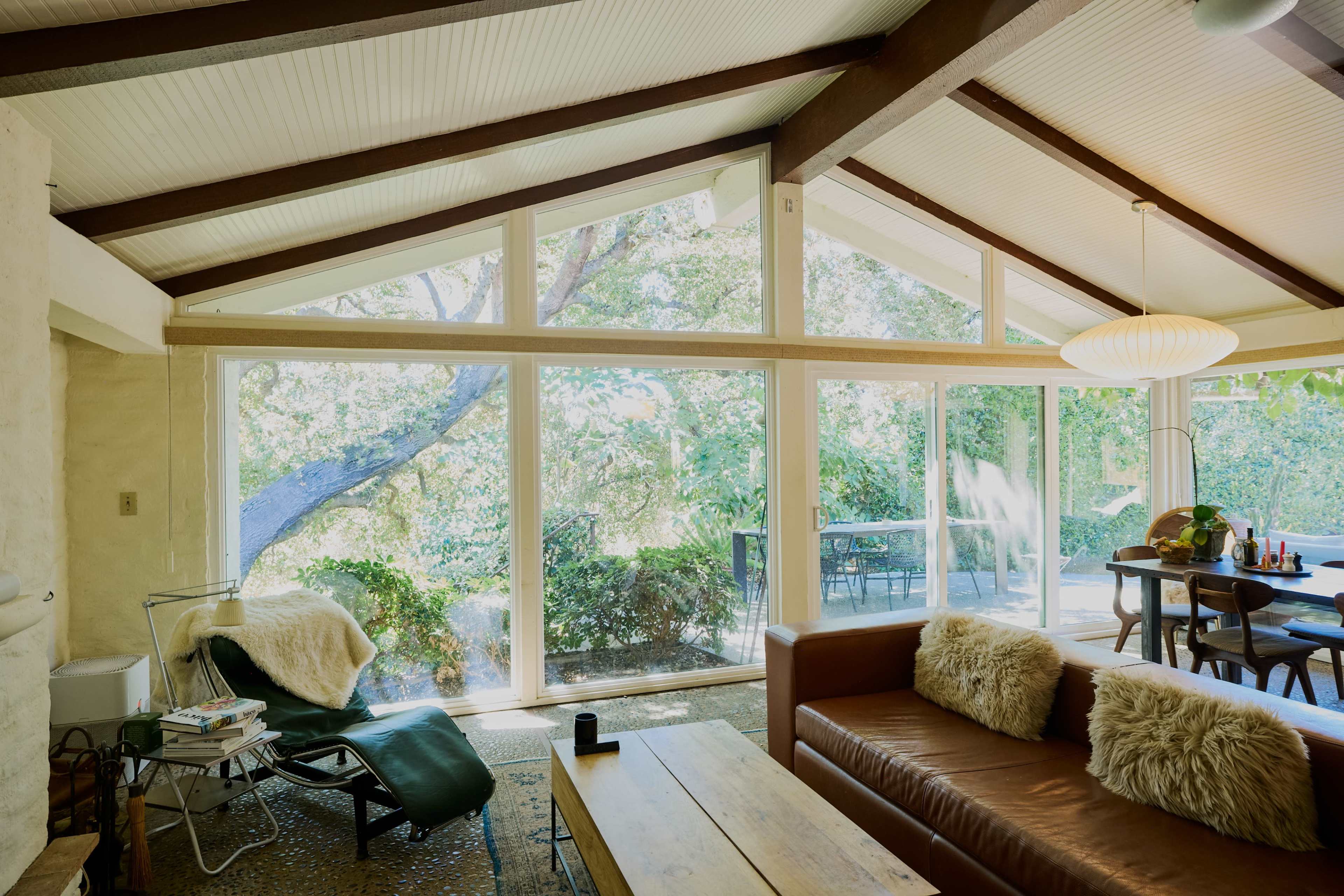 A modern living room with large windows that overlook a garden, featuring a brown sofa, a wooden coffee table, and a dining area in the background.