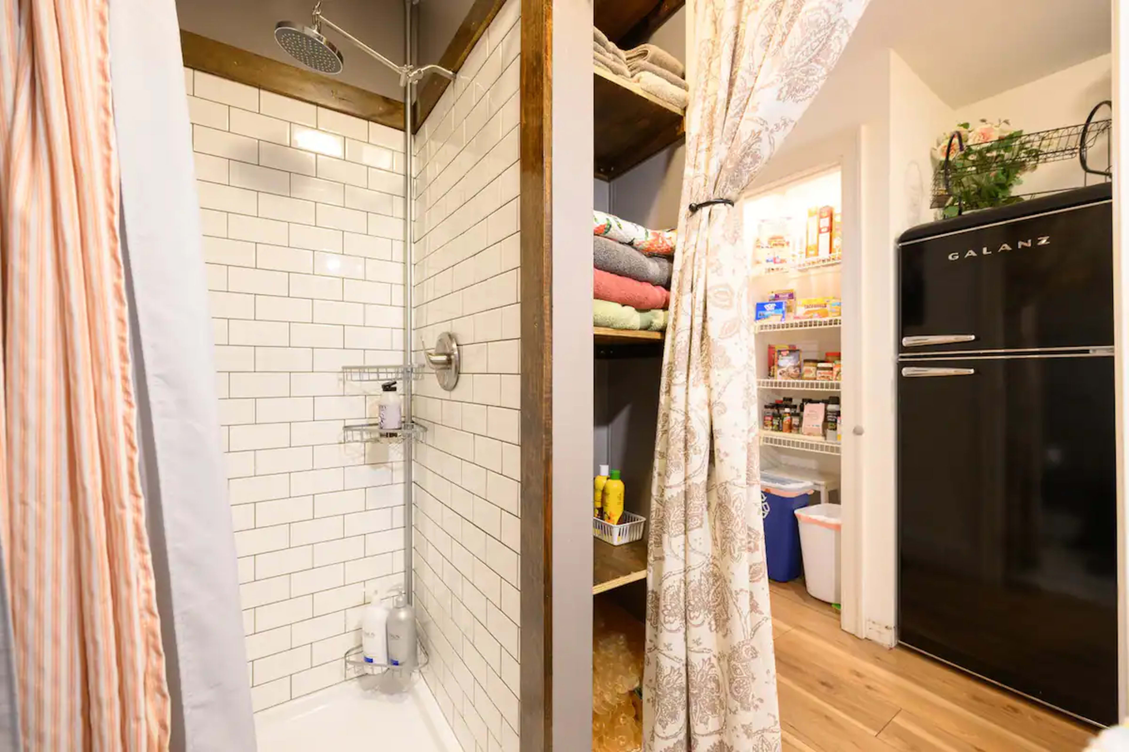 A shower area with white tiled walls is separated by a curtain from a small pantry equipped with shelves and a black refrigerator.