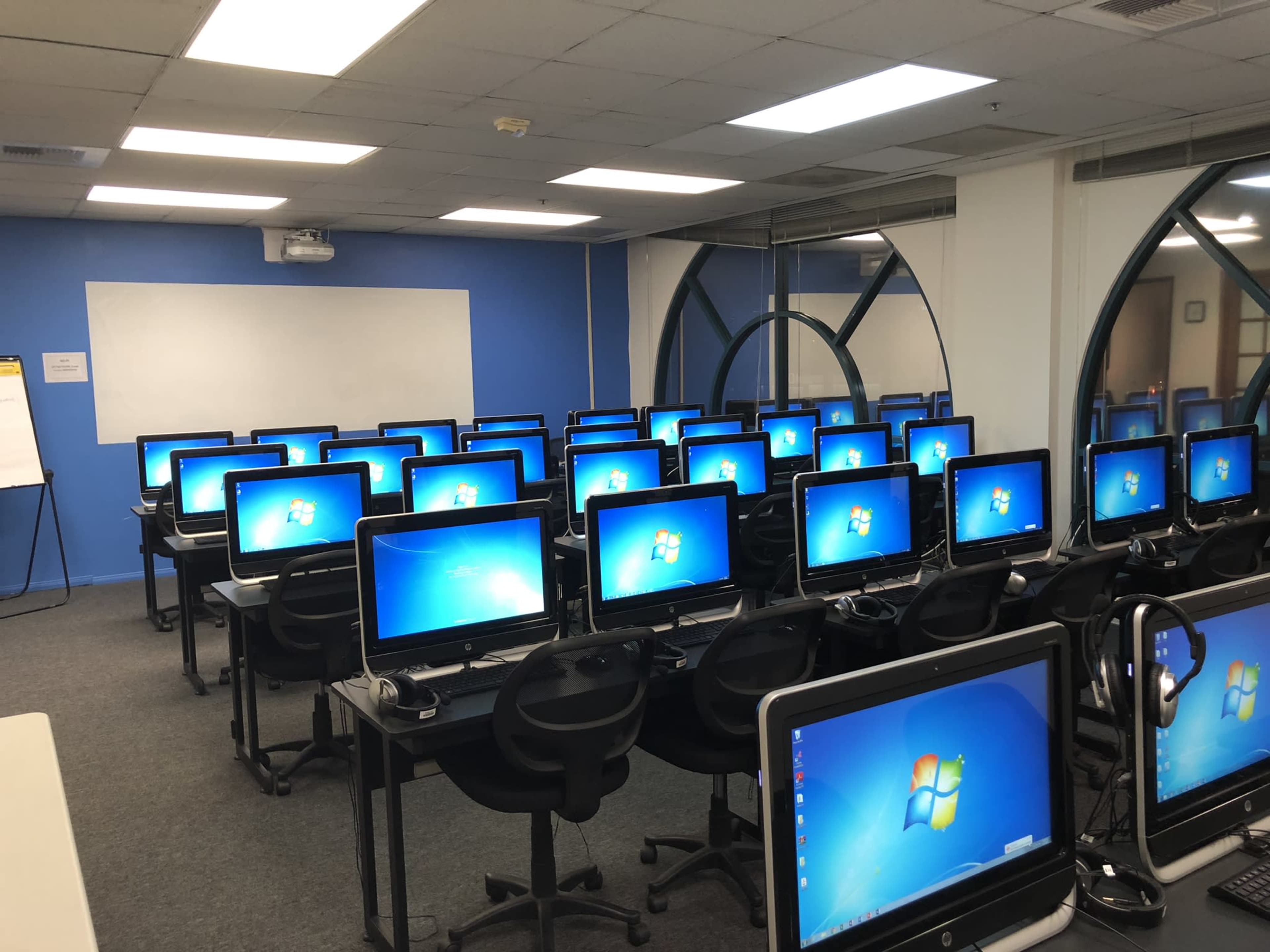 A computer lab with multiple rows of desktop computers facing a whiteboard and a projector screen, all set against a blue wall.