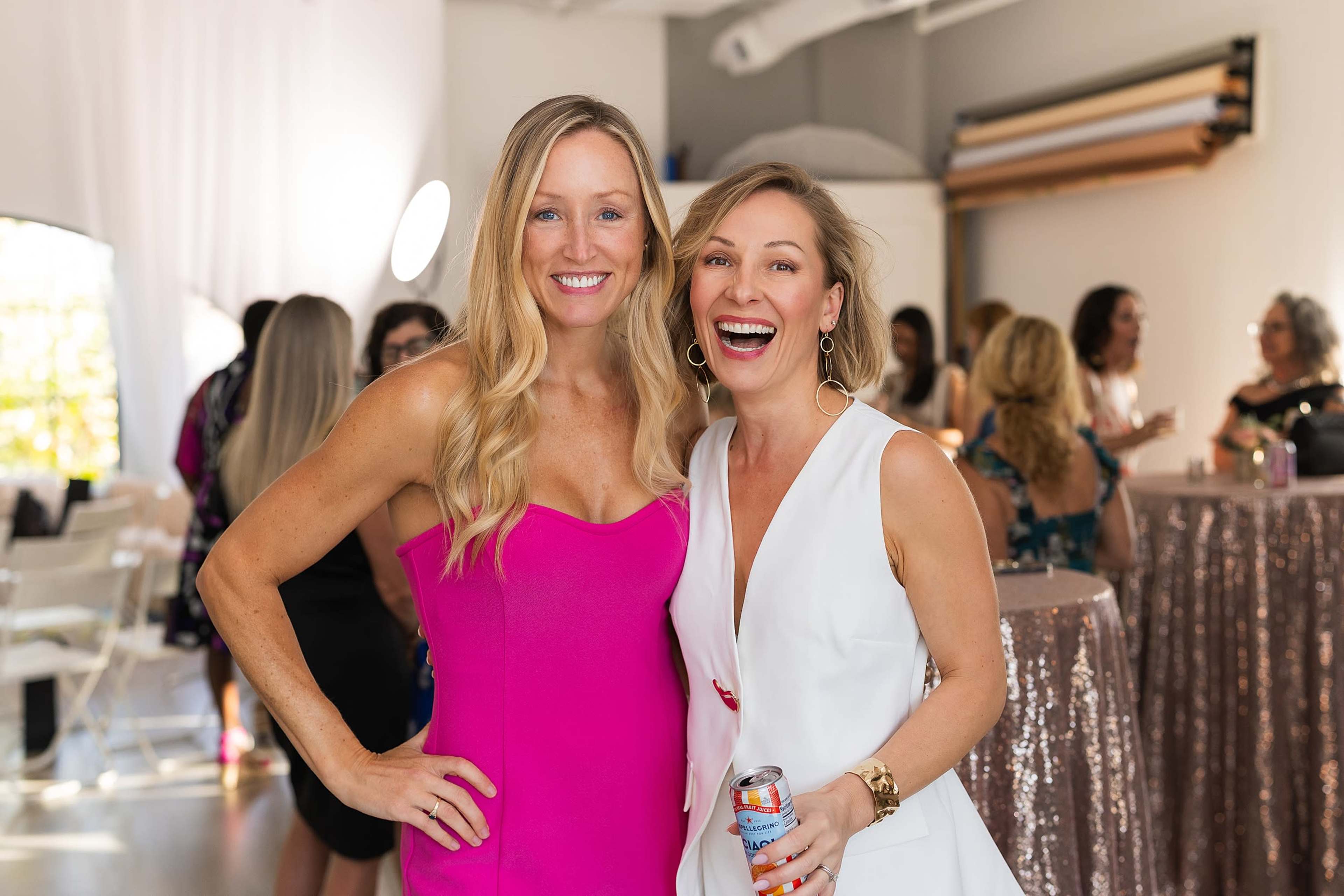 Two women pose together for a photo in a bright indoor setting, surrounded by other people and elegantly decorated tables.