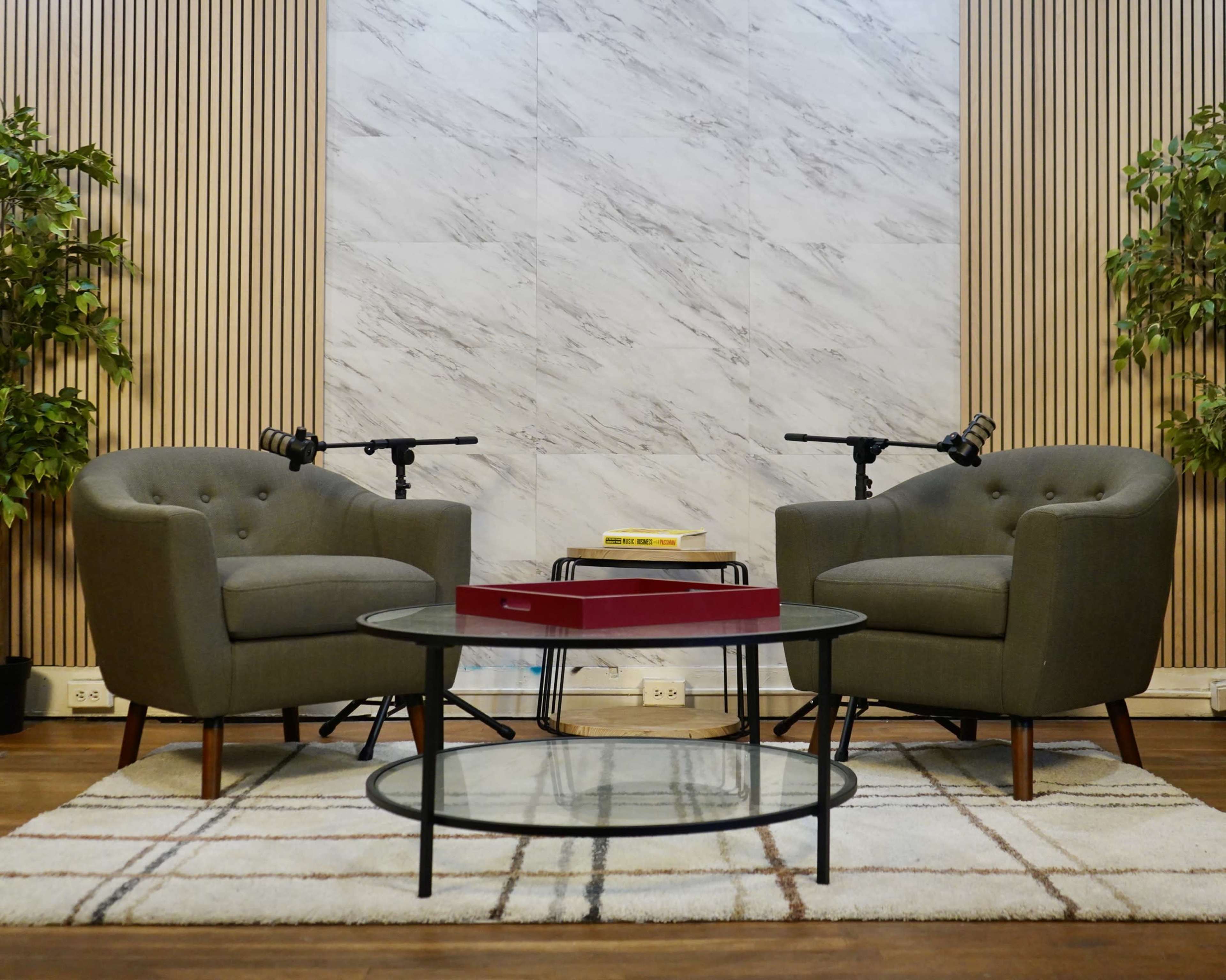 Two gray armchairs are positioned facing each other around a circular glass coffee table, with a red tray on it, against a marble wall adorned with greenery.