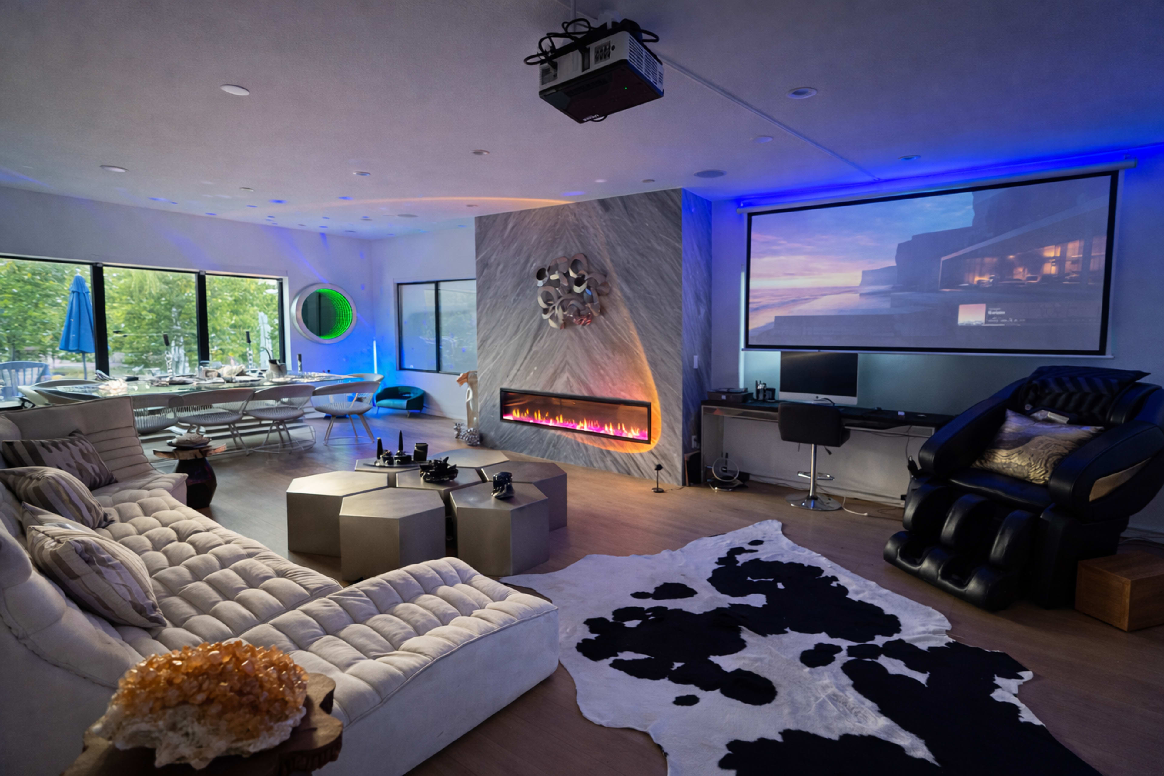 The image shows a modern living space featuring a plush sofa, a sleek fireplace, a large projector screen, and a dining area with large windows.