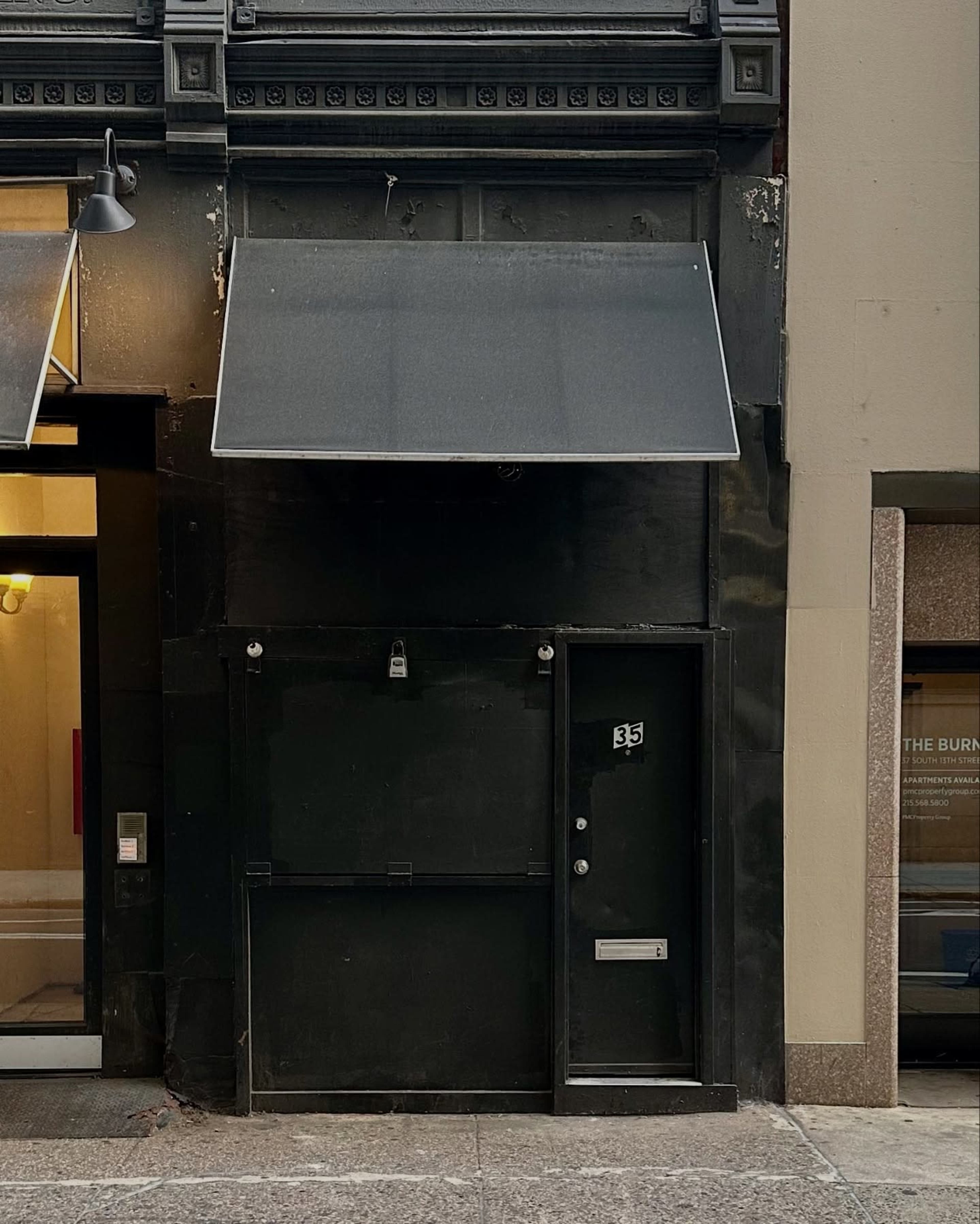 The image shows a dark exterior wall of a building featuring a black awning and a door with a small window.