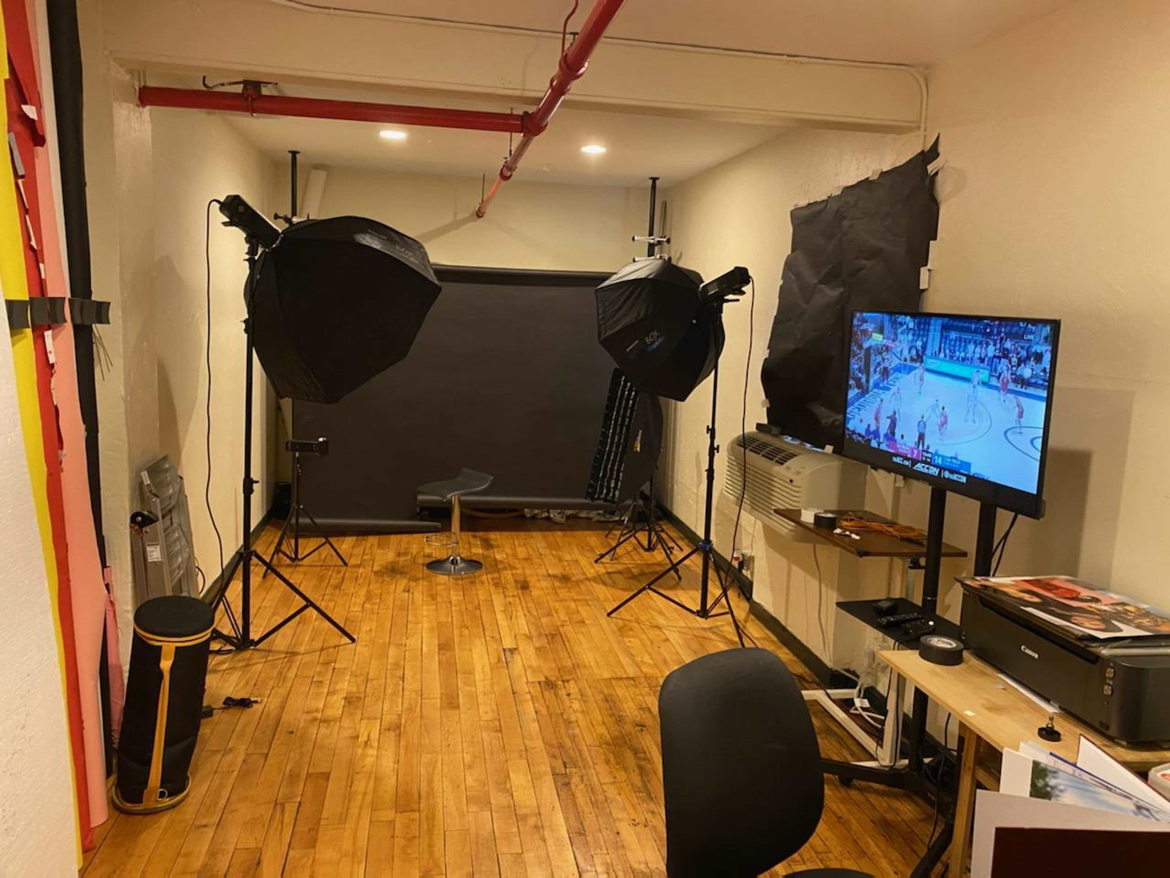 The image shows a small studio space with two softbox lights, a black backdrop, a TV displaying a basketball game, and a desk with a chair.