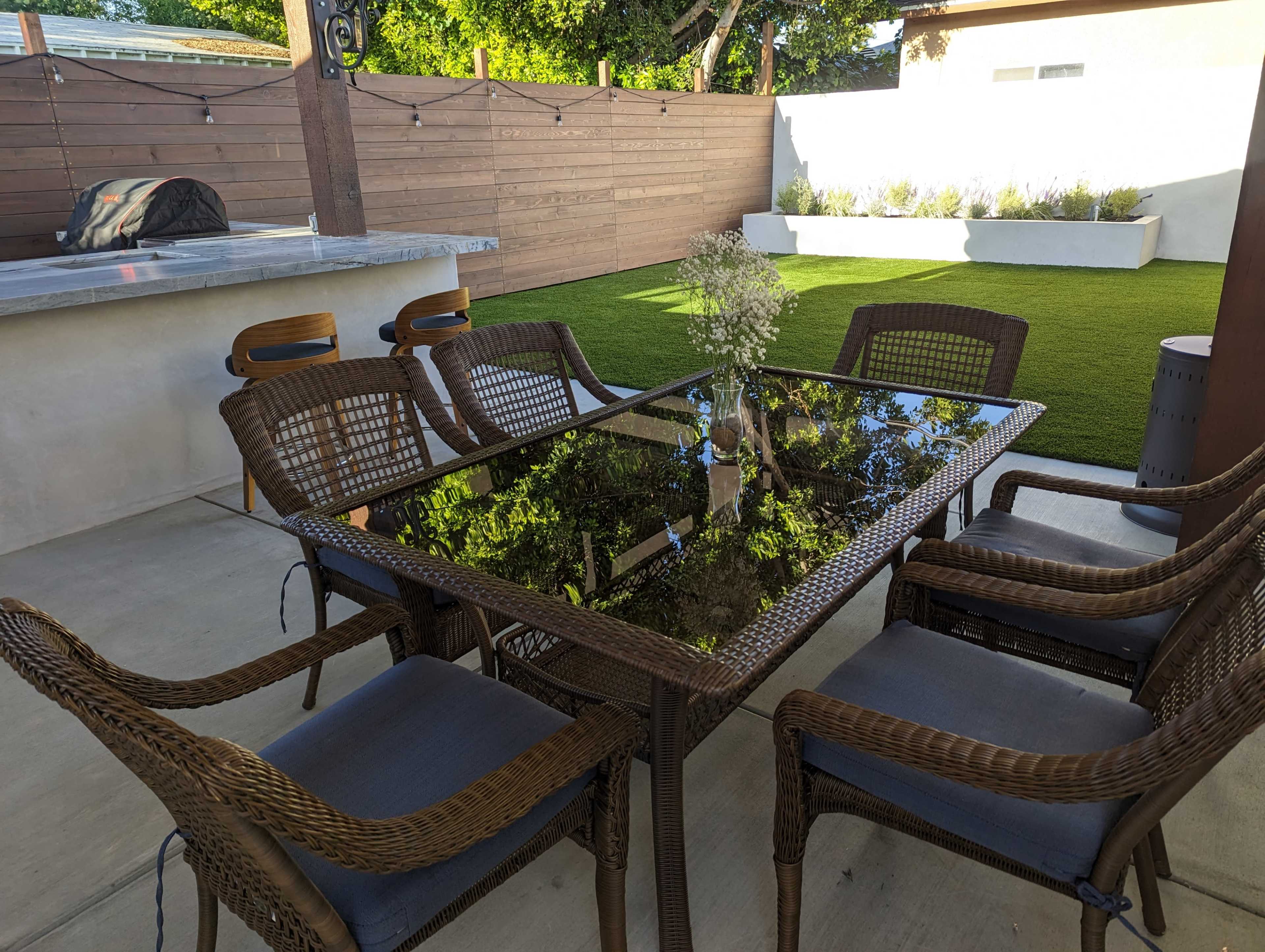 A glass-topped dining table surrounded by six wicker chairs is set on a concrete patio, with a grassy area and wooden fence in the background.