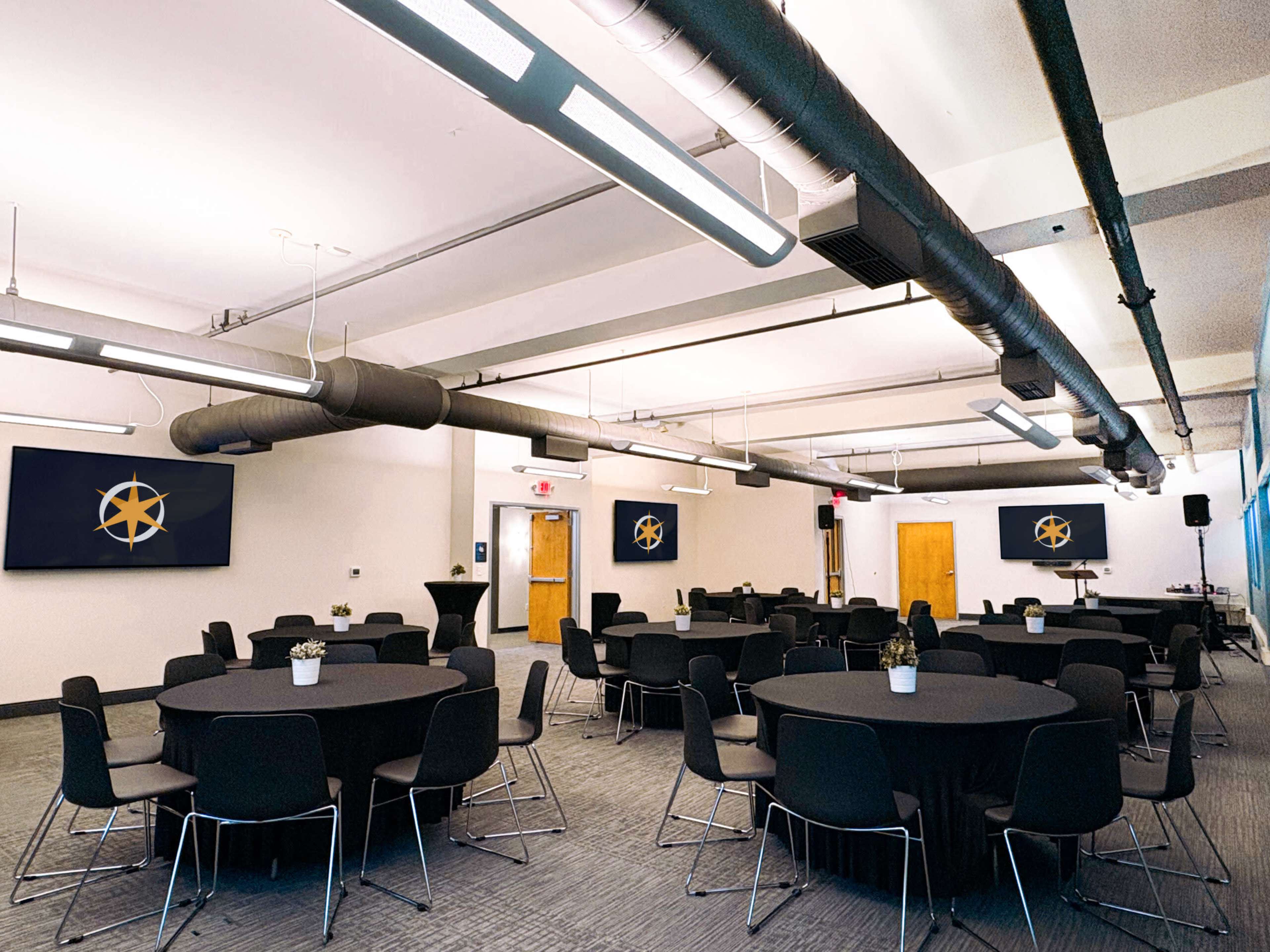 The image shows a spacious meeting room set up with black round tables and chairs, featuring two large screens on the walls displaying a logo.