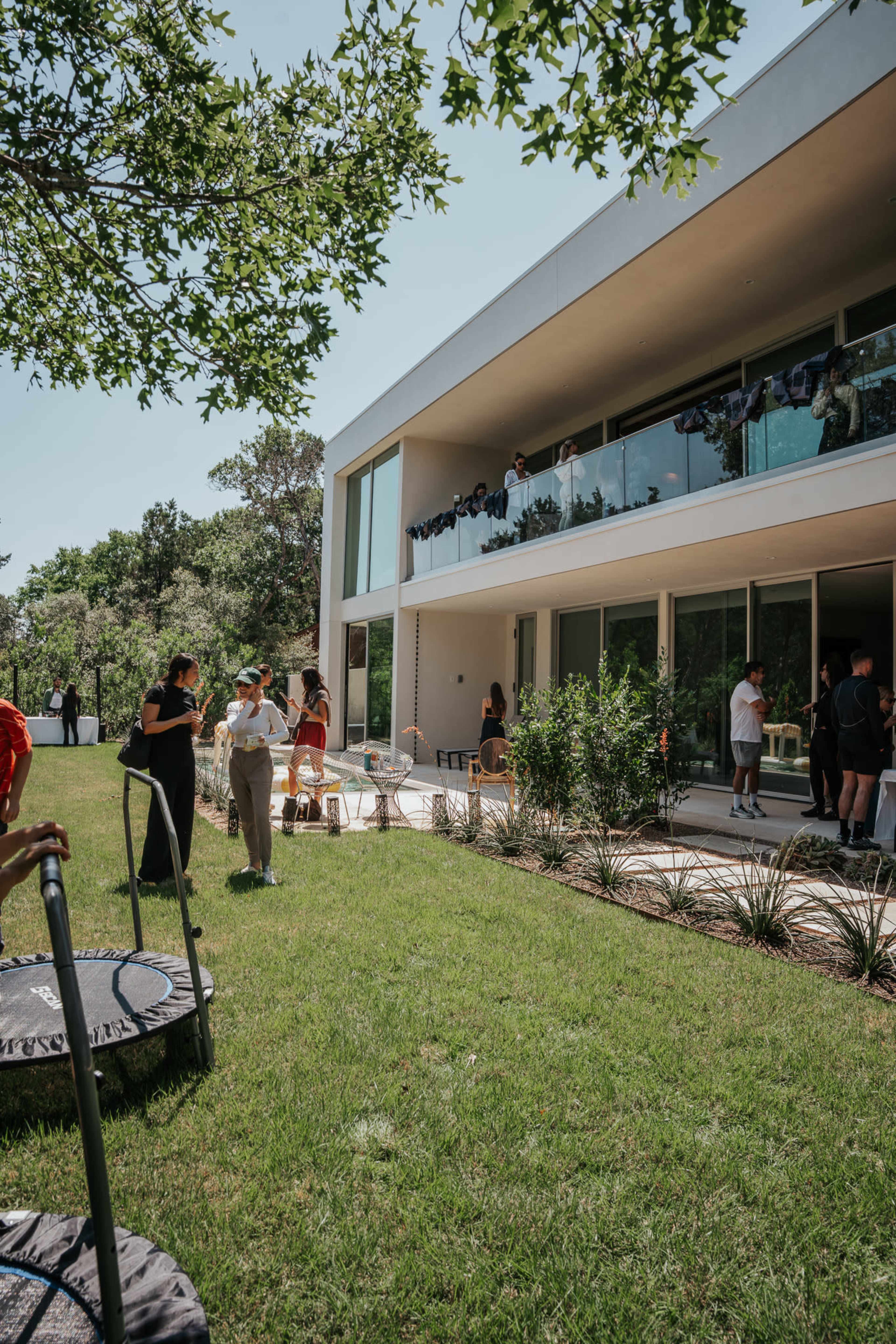 A modern building with large glass windows overlooks a grassy area where several people are engaged in conversation.