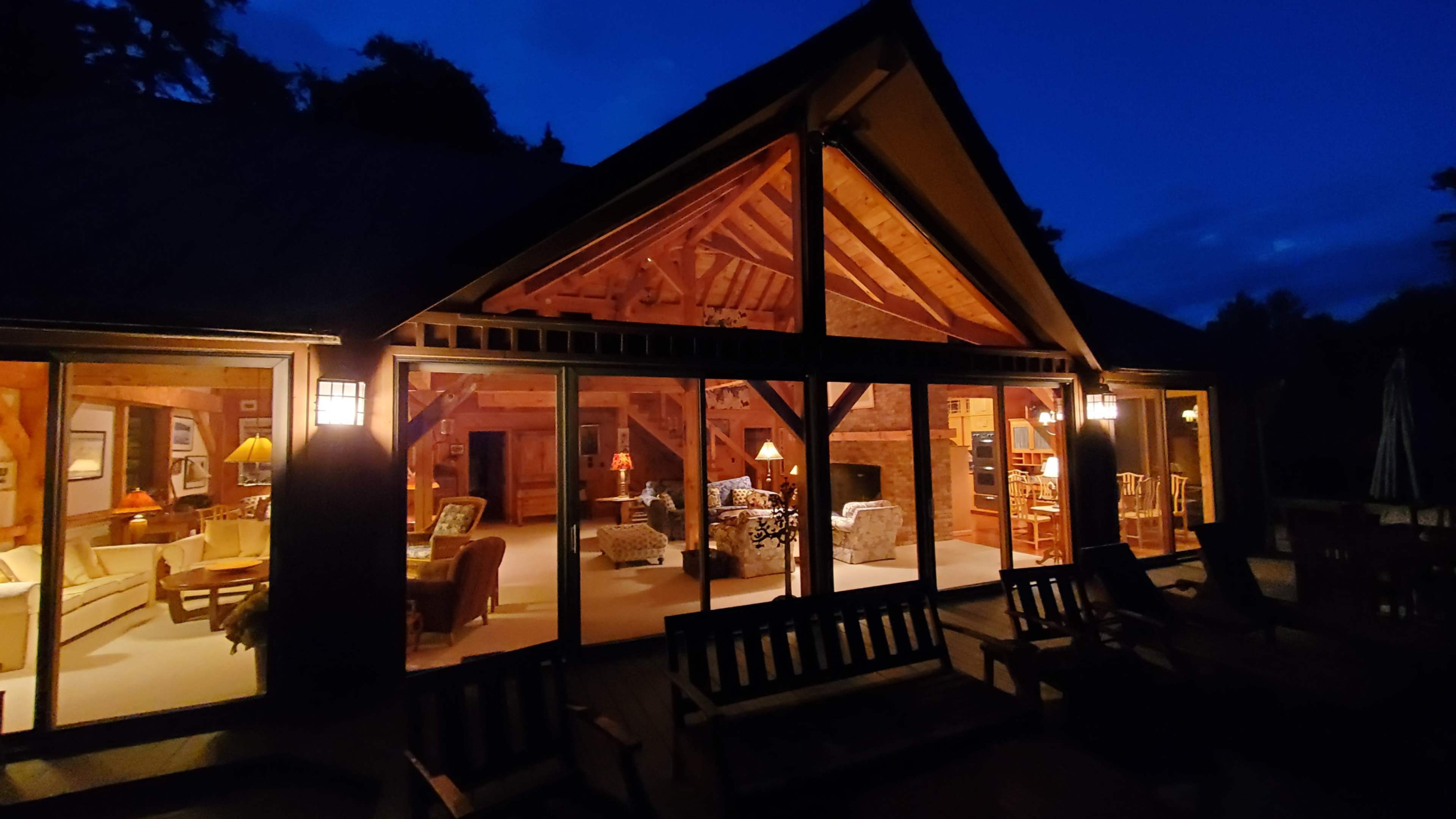 A timber-framed house is illuminated at night, showcasing a cozy interior through large glass windows.