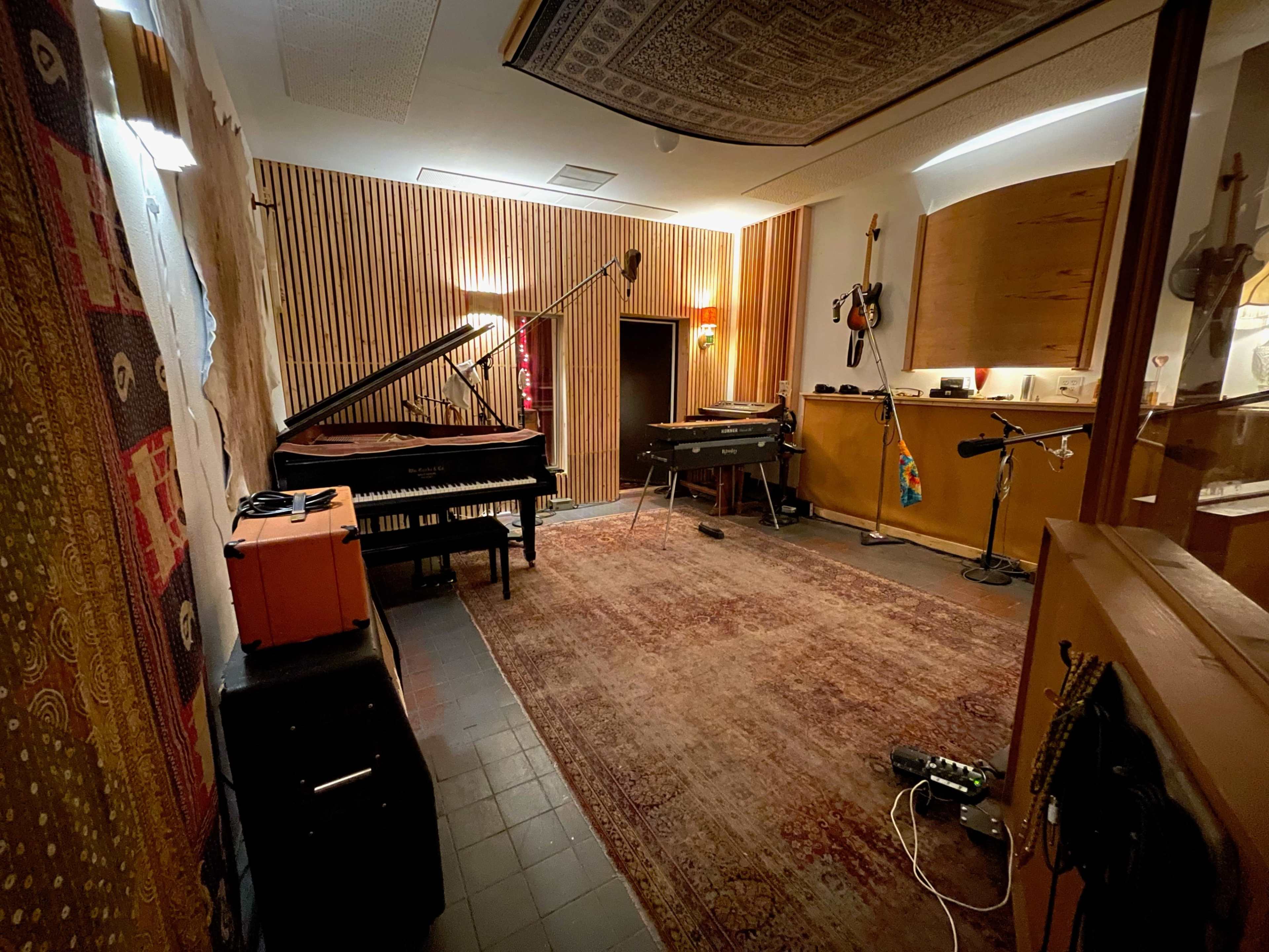 A music recording studio featuring a grand piano, several electric keyboards, microphones on stands, and sound-absorbing wooden panels on the walls.