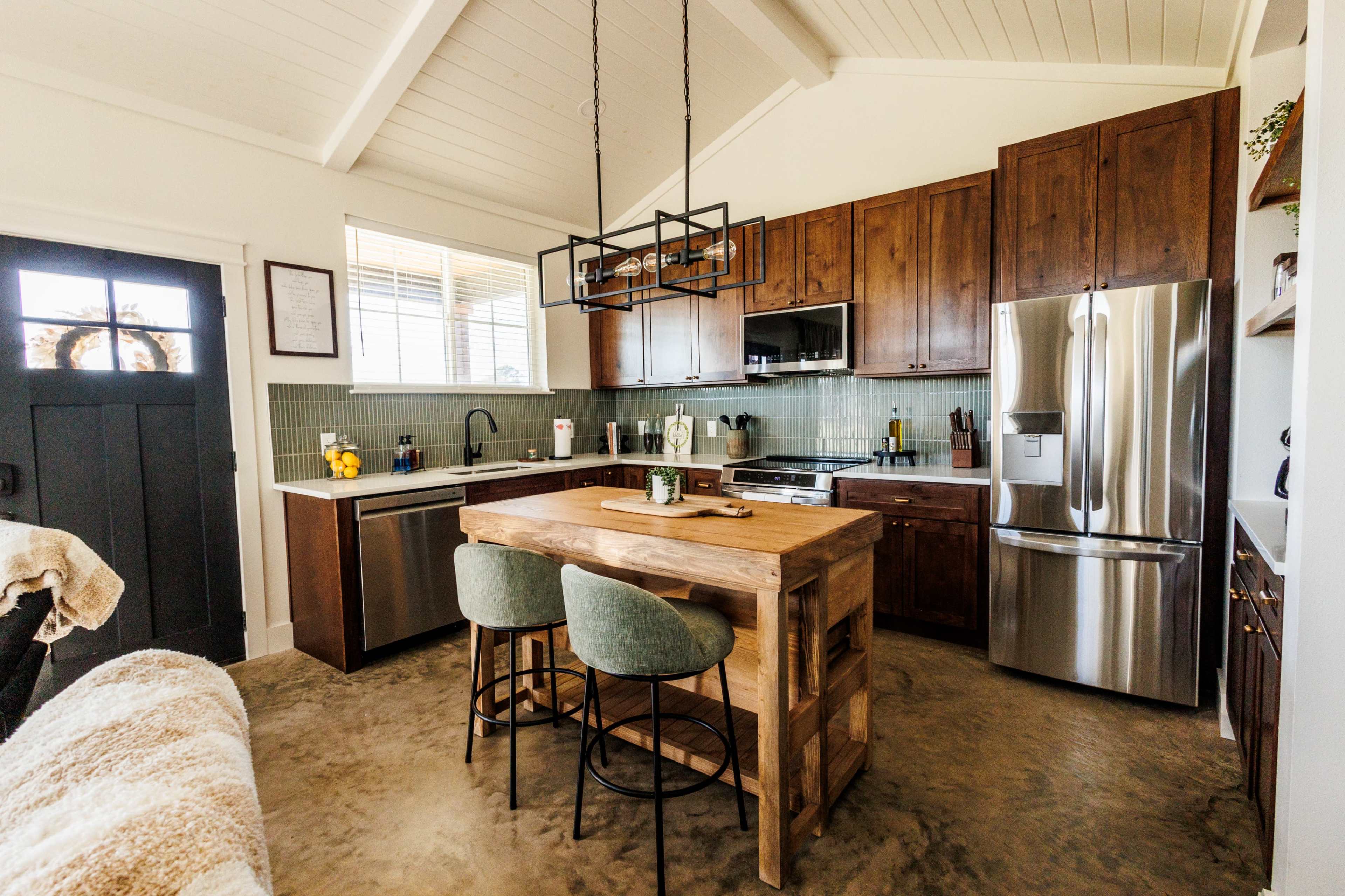 The image shows a modern kitchen with wooden cabinets, stainless steel appliances, and a central wooden island with two stools.