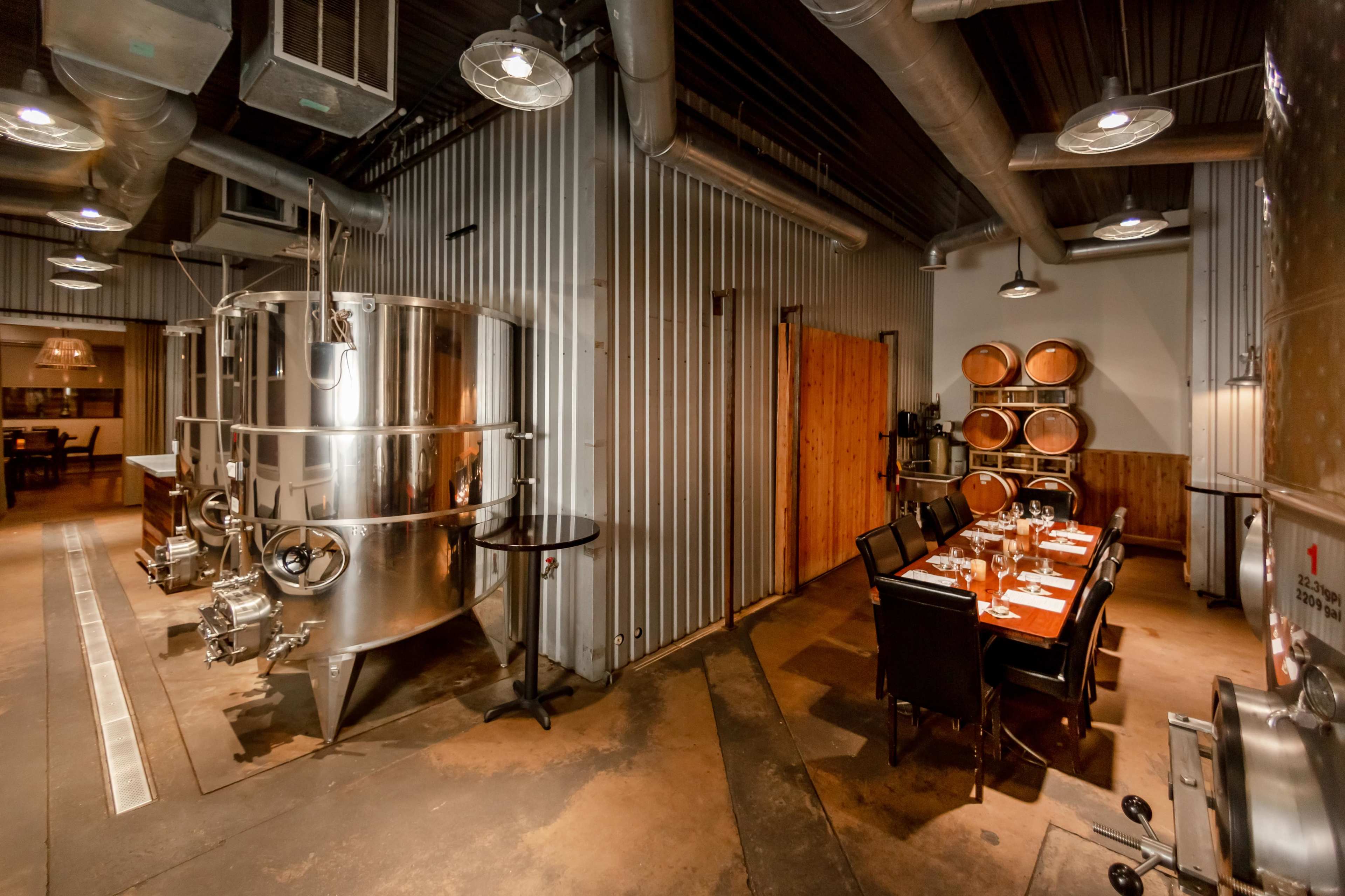 A winery interior featuring stainless steel fermentation tanks, wooden barrels, and a dining table set for a meal.