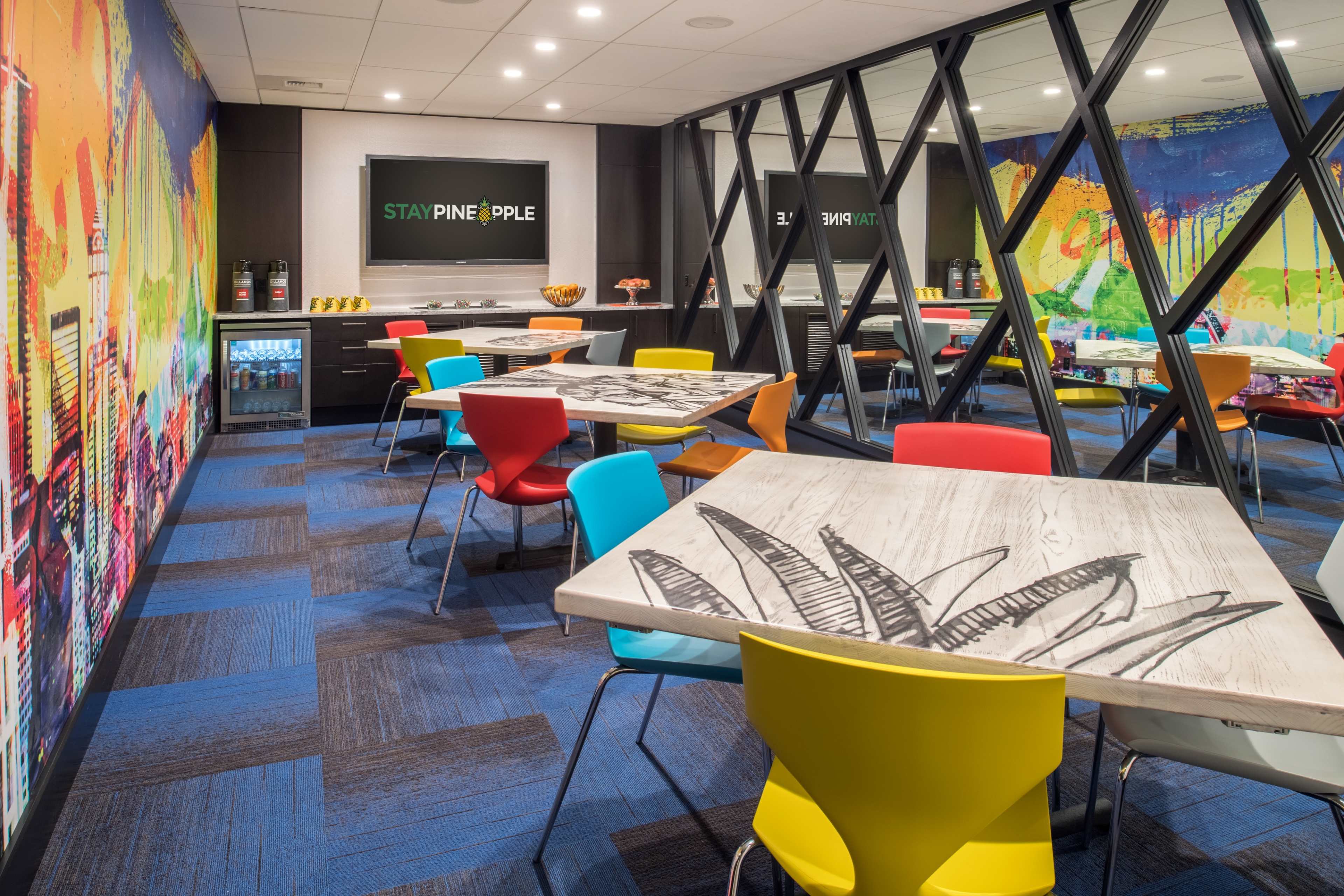 A colorful break room with tables and chairs arranged around a central area, featuring large windows and an artistic mural on the walls.