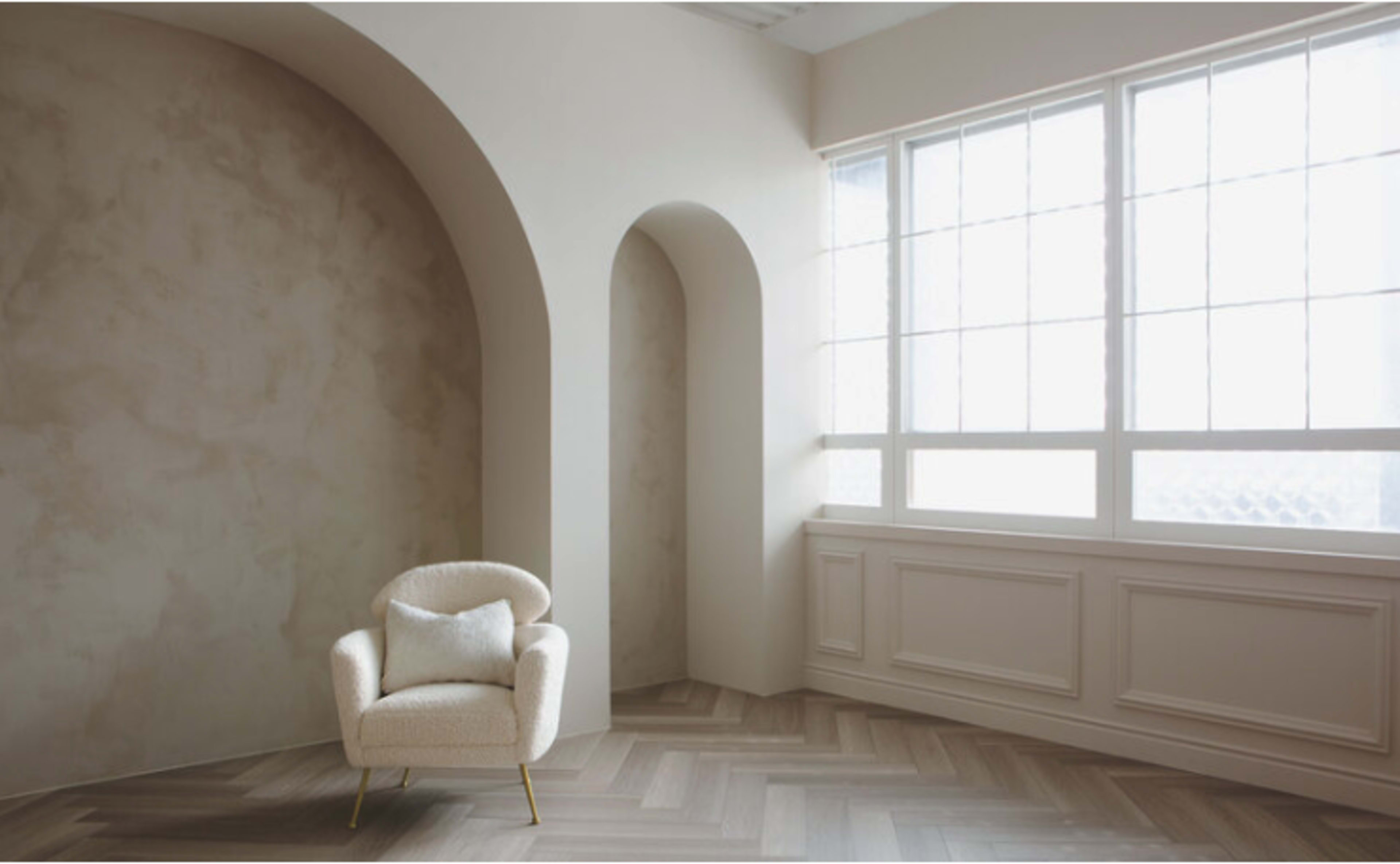 A light-filled room features a minimalist upholstered chair positioned against a textured wall and arched alcove near a large window.