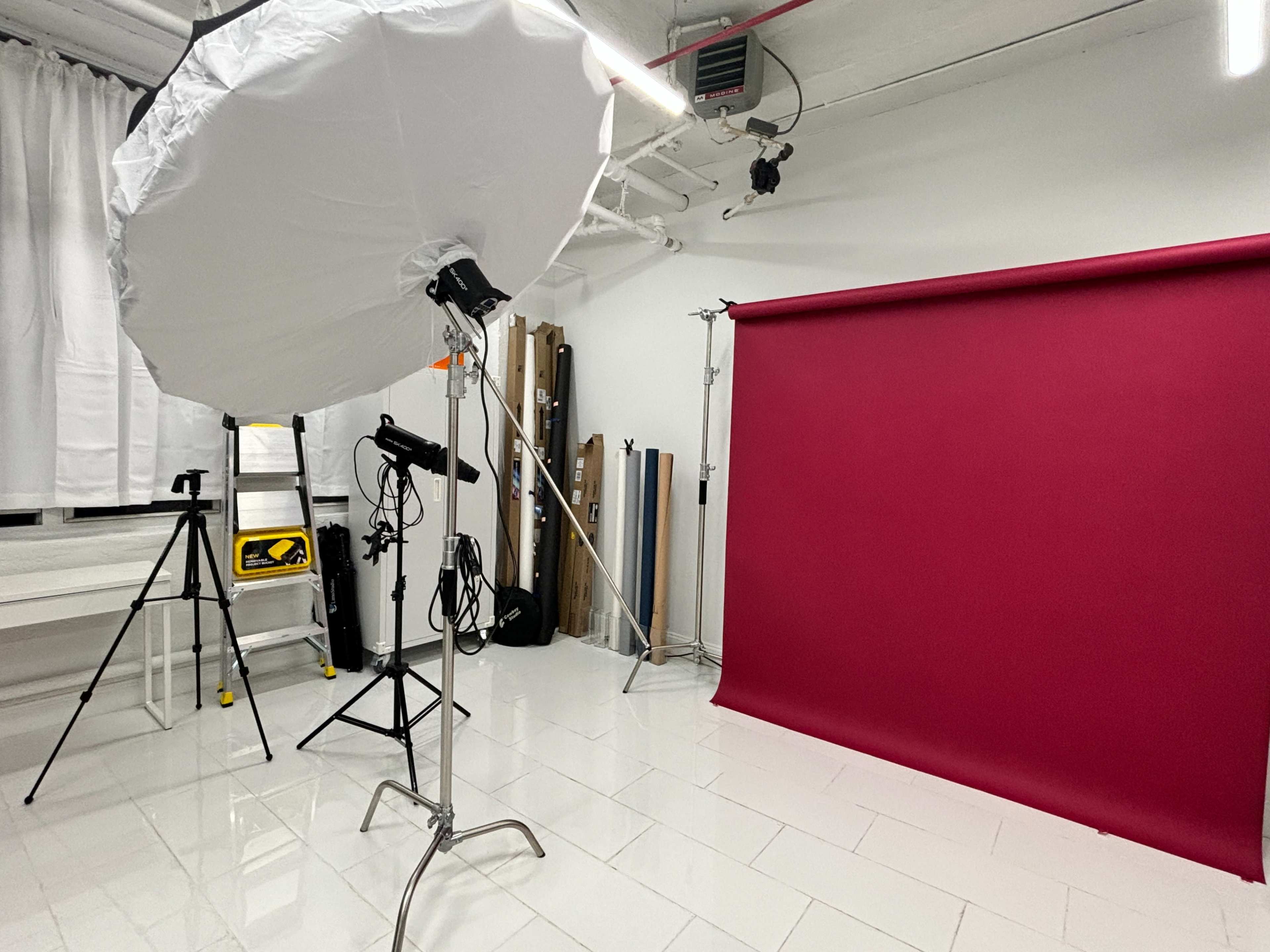 The image shows a photography studio setup with a large softbox light, a tripod, and a solid burgundy backdrop against a white tiled floor.