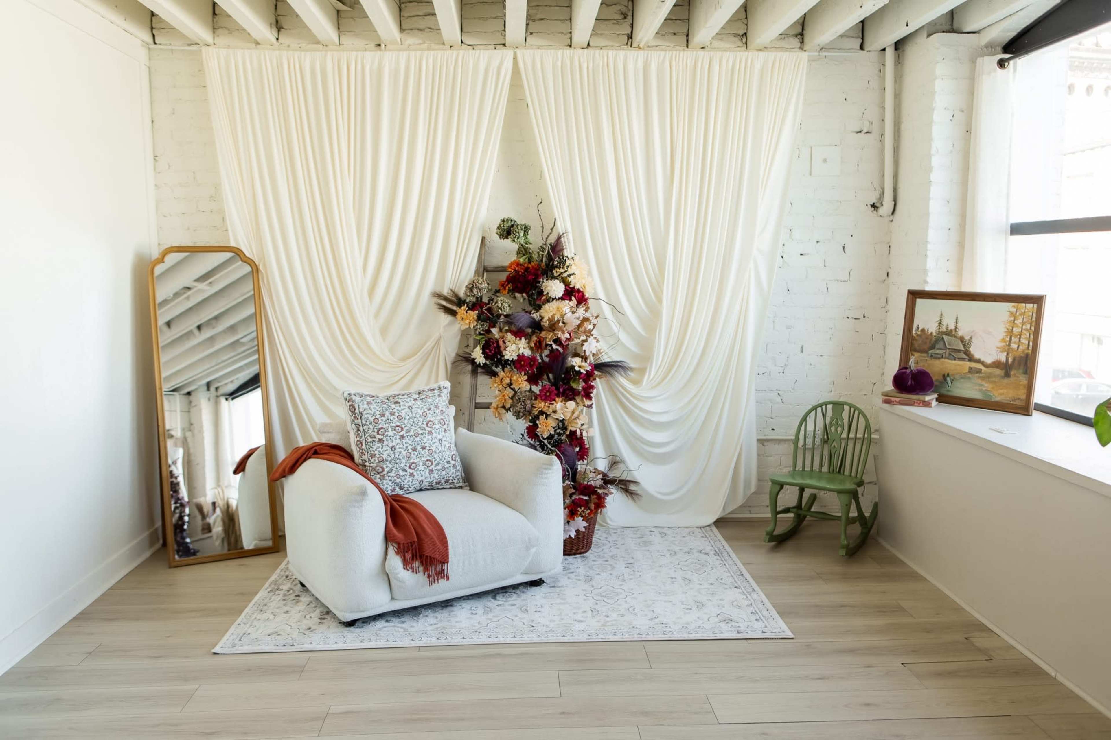 The image shows a cozy interior scene featuring a white armchair, a large mirror, a floral arrangement, and a small green chair against a backdrop of white drapes and light-colored flooring.