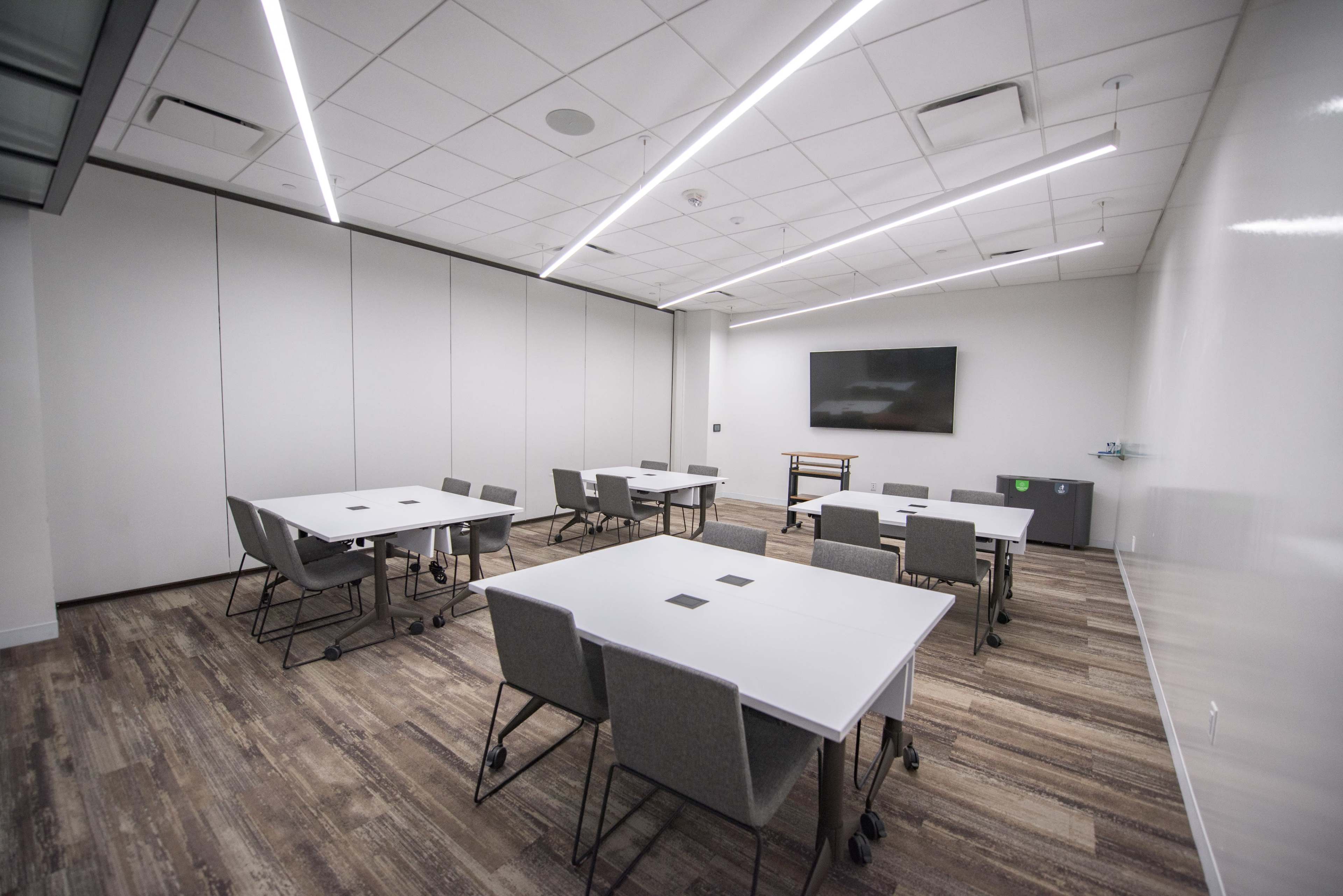 A modern conference room features multiple white tables and gray chairs arranged around the space, with a wall-mounted screen at one end.