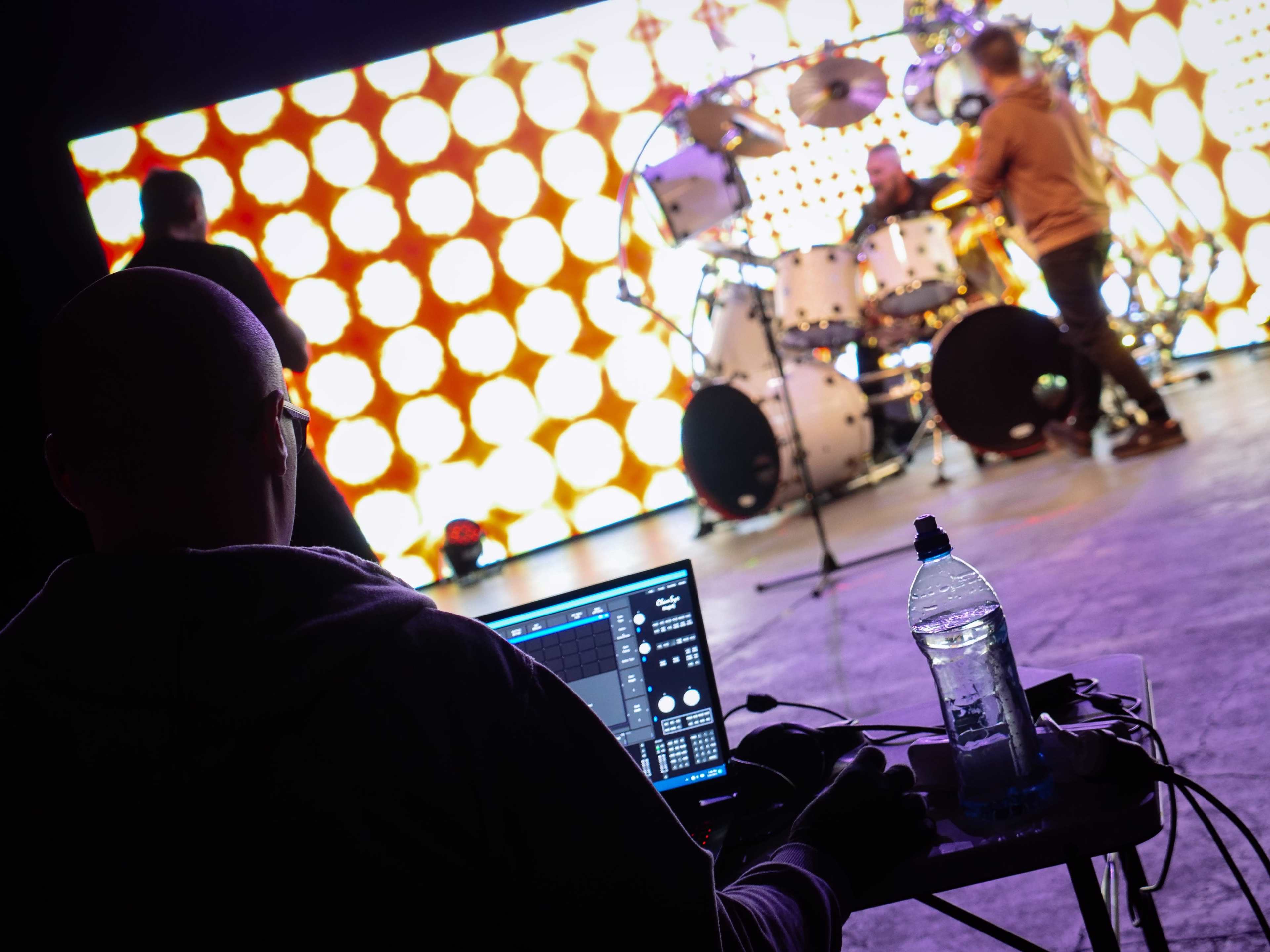 A person sits at a laptop in the foreground, while a drummer plays on stage in front of a backdrop of bright, circular lights.