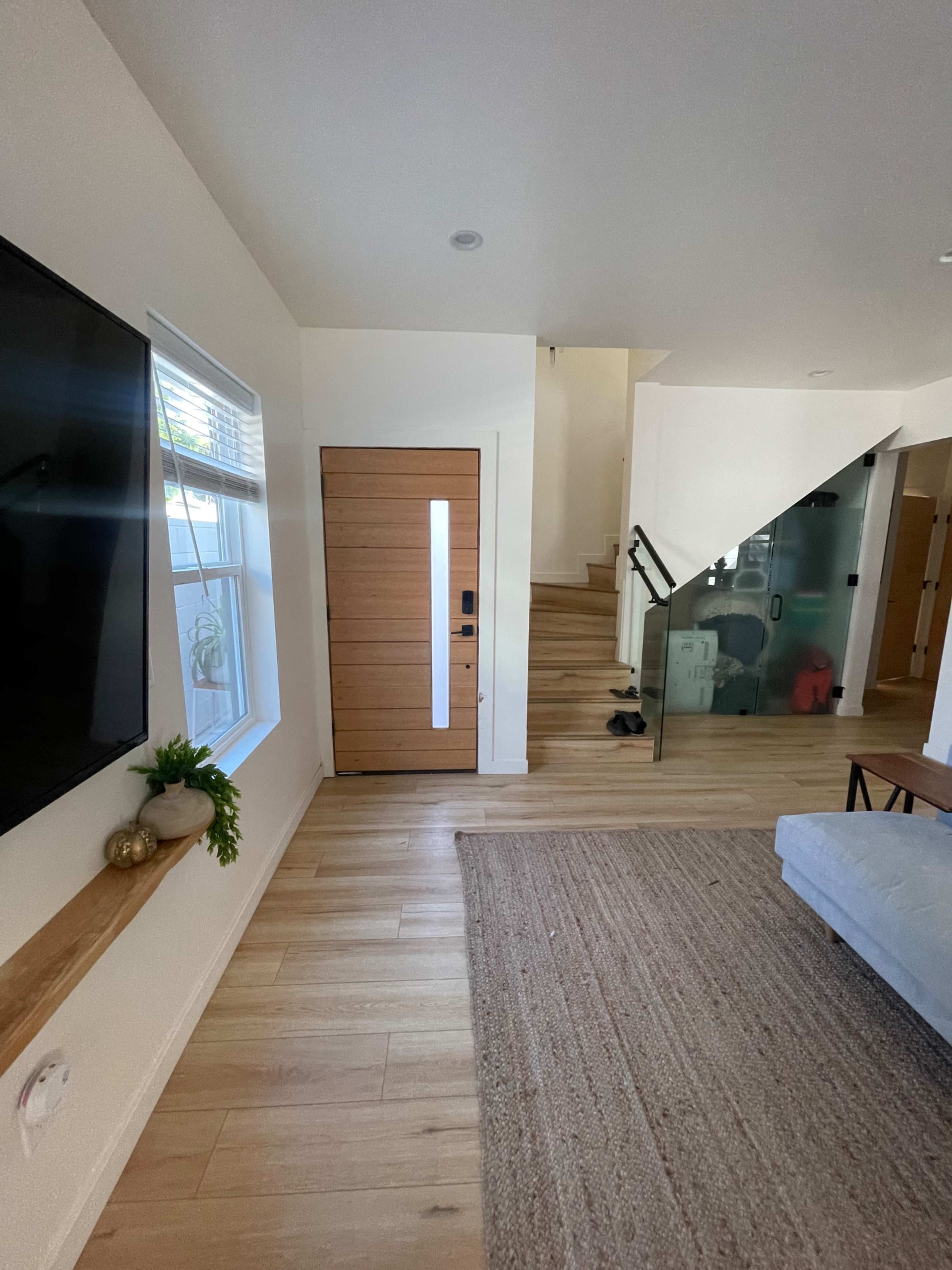 A modern entryway with a wooden front door, a television mounted on the wall, and a staircase leading to an upper level.