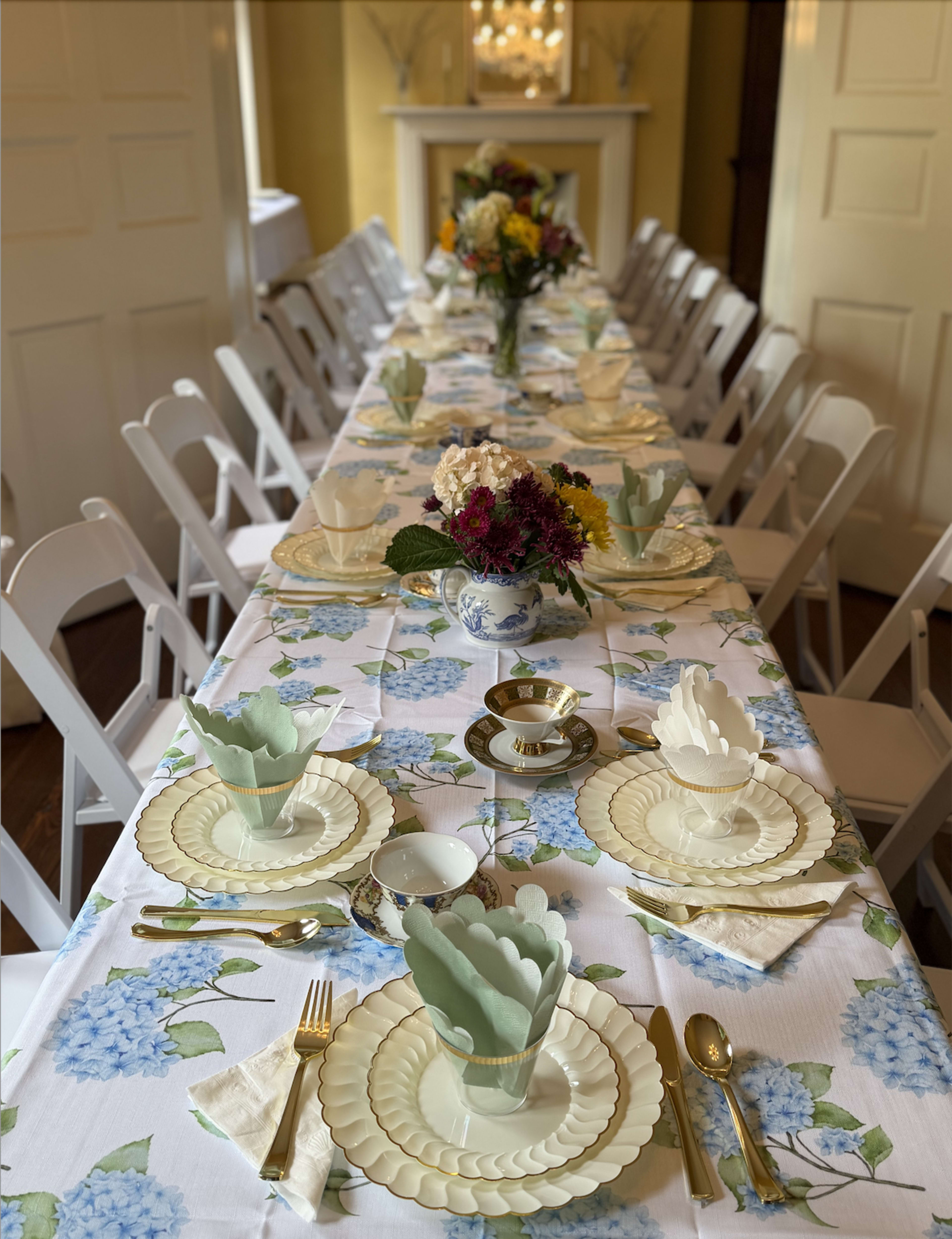 A long dining table is set with floral-patterned tablecloth, matching dinnerware, and decorative utensils, surrounded by white folding chairs.