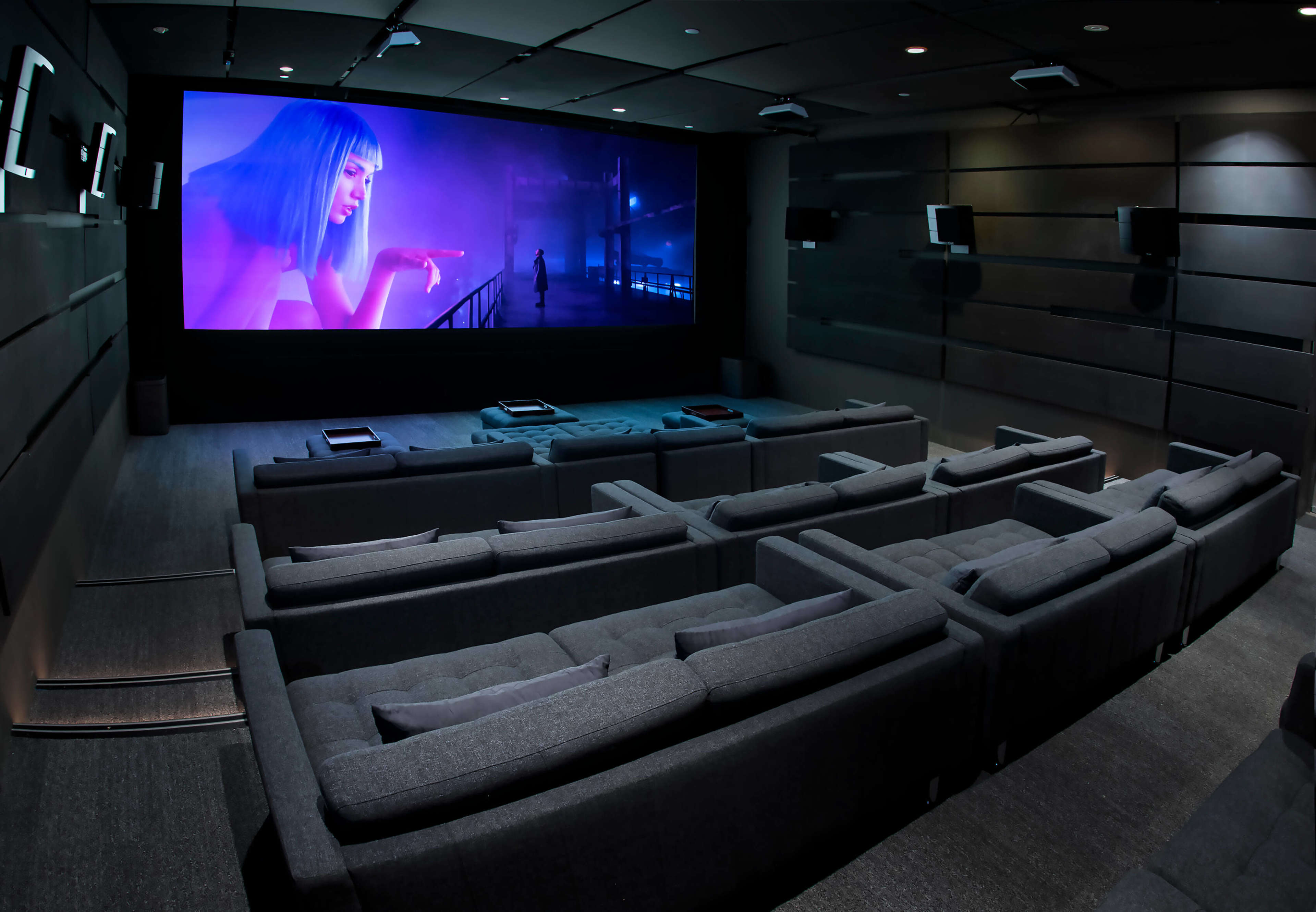 The image shows a modern home theater with multiple gray couches arranged in a tiered seating layout facing a large screen that displays a colorful animated scene.