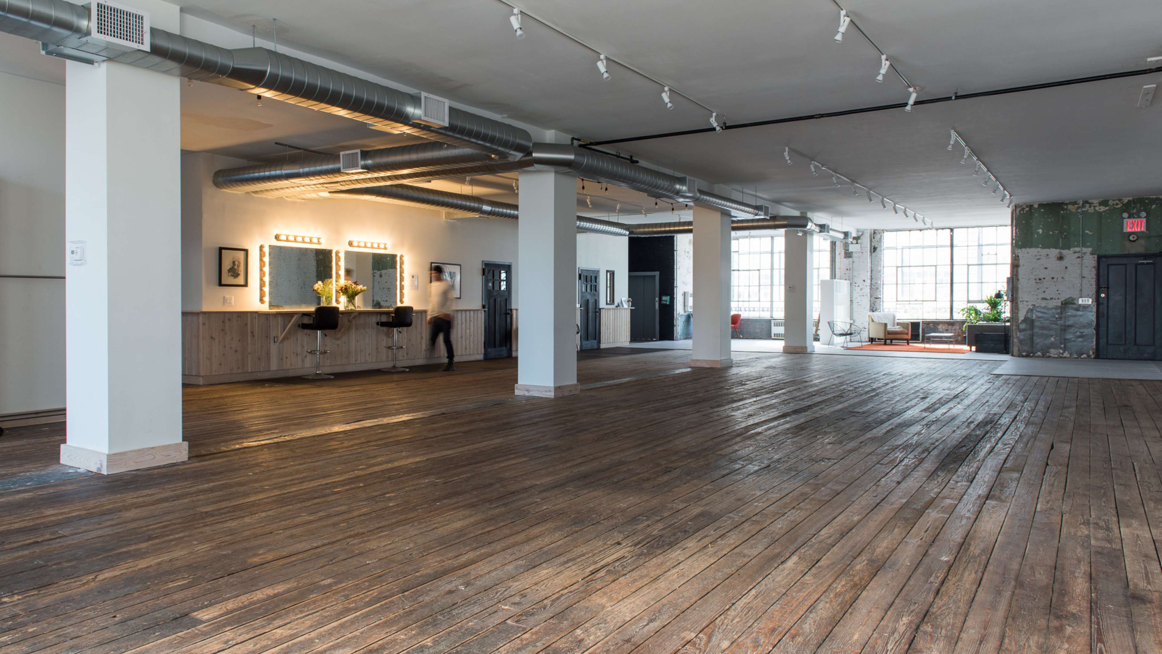 The image shows a spacious interior with wooden floors, large windows, and minimalist decor featuring a few furnishings and fixtures along the walls.