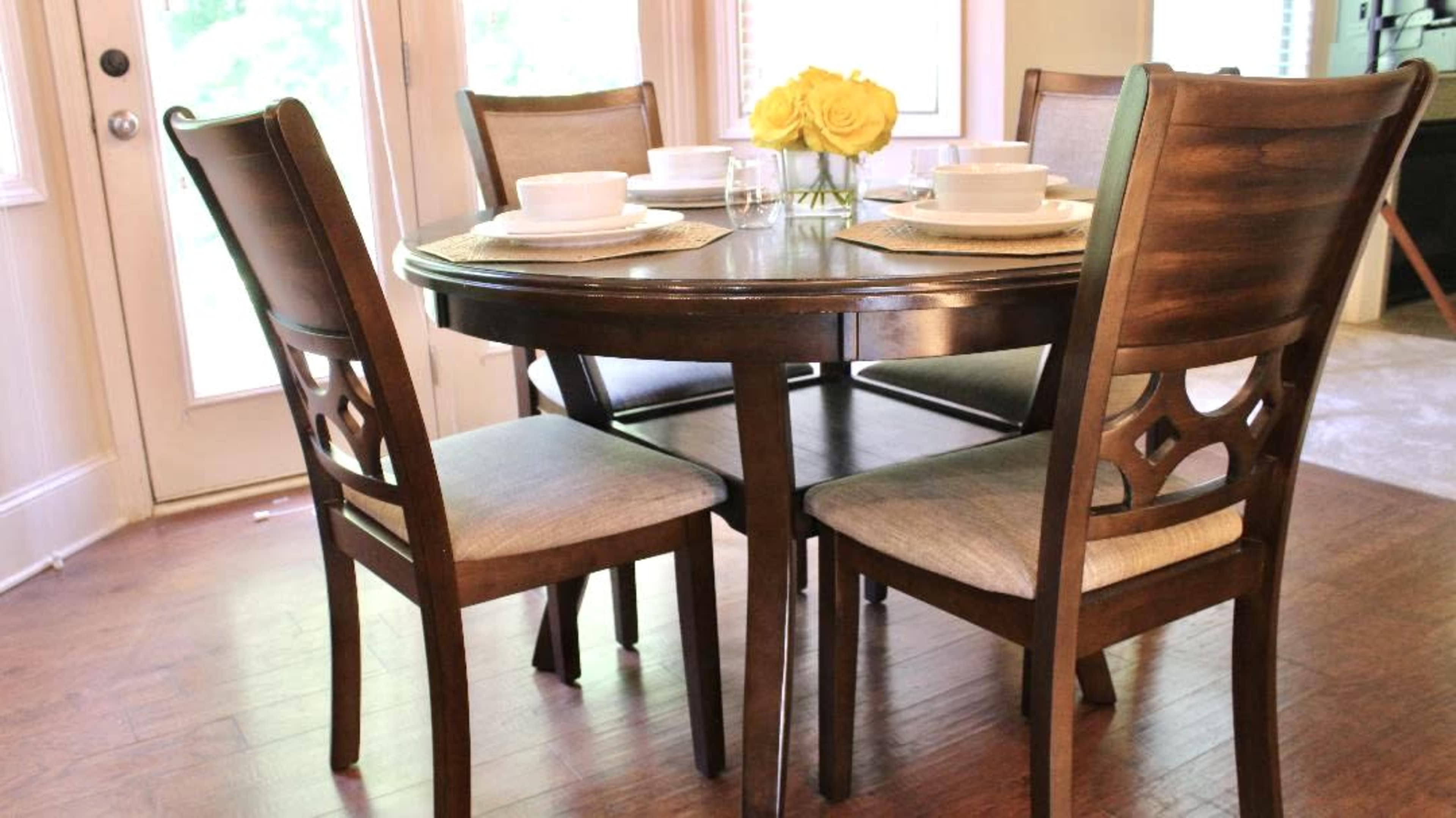 A round wooden dining table is set with white dishes and a vase of yellow roses, surrounded by four chairs, in a brightly lit room.