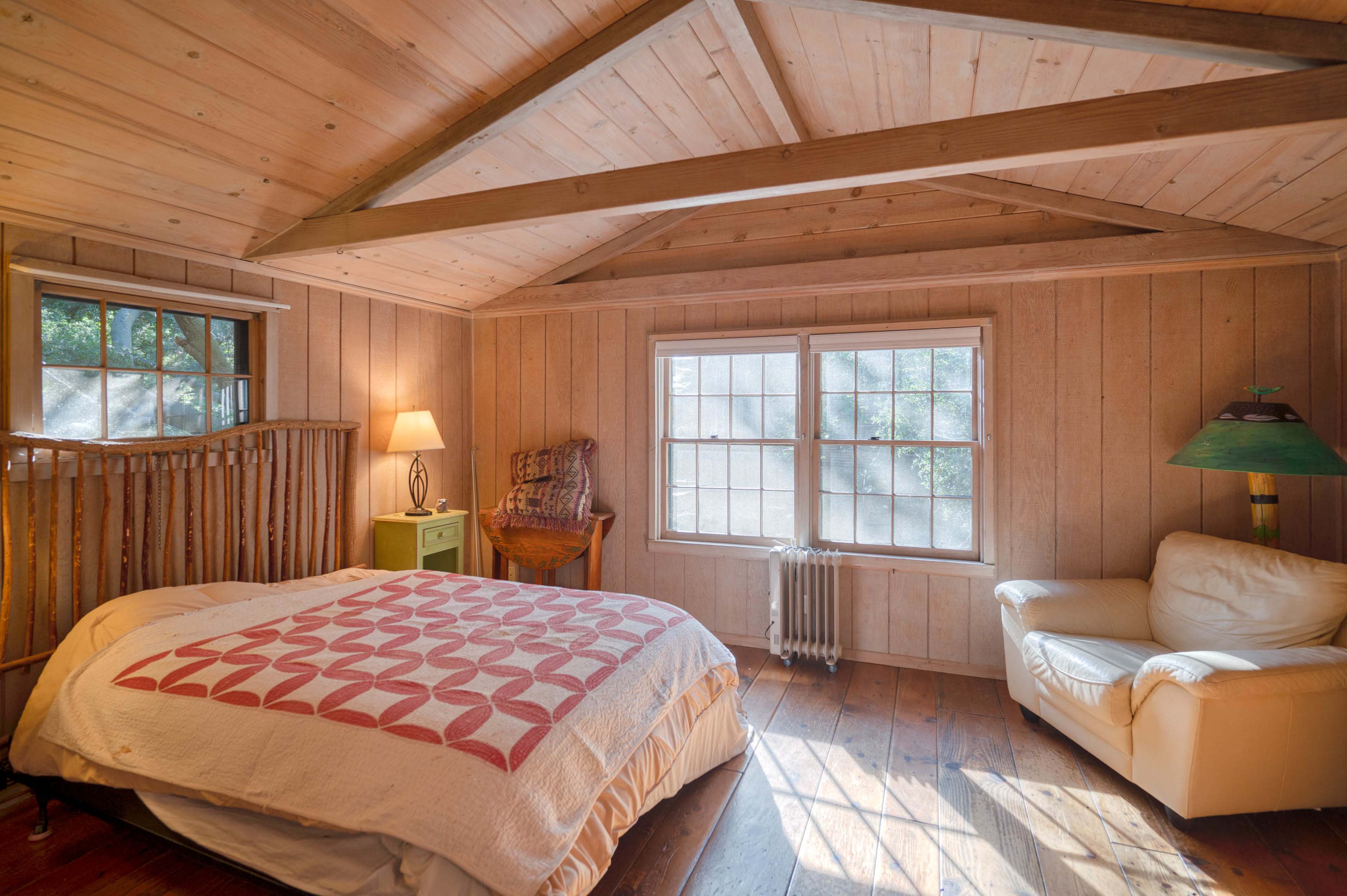 The image shows a rustic bedroom featuring wooden walls, a bed with a quilt, a chair, a lamp, and large windows allowing natural light.