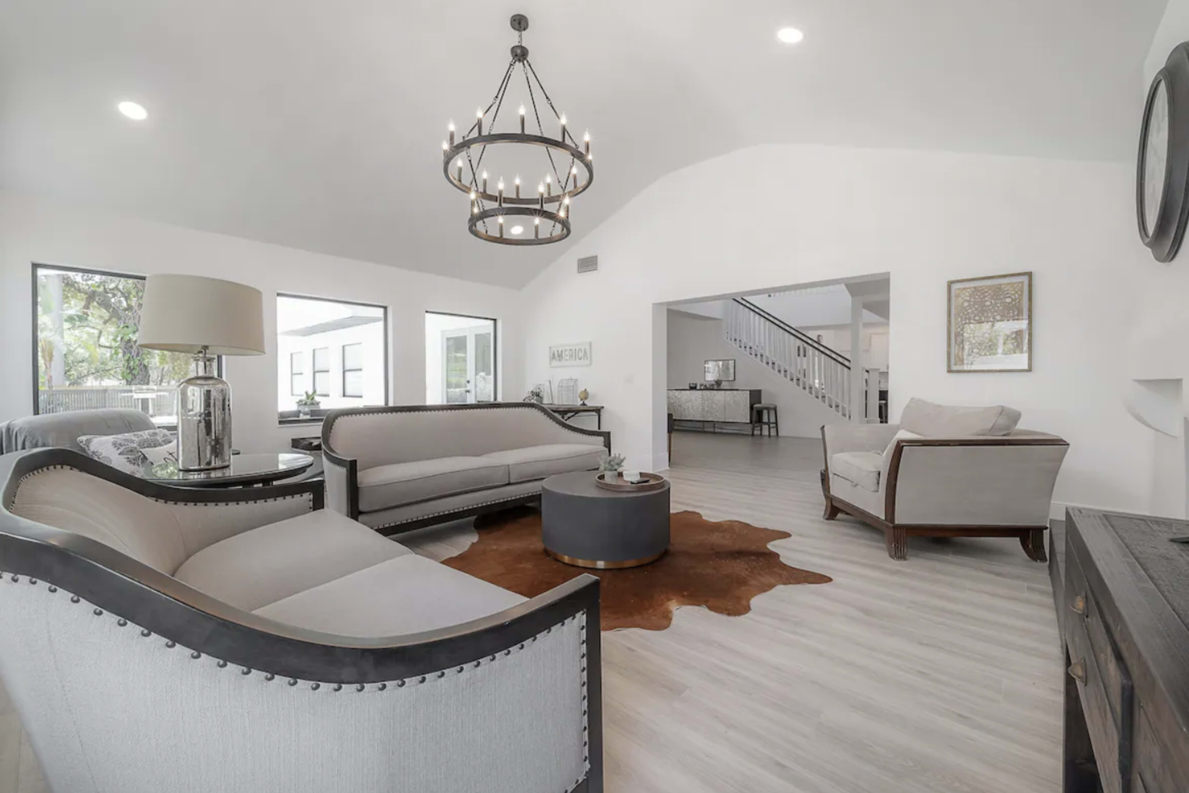 The image shows a spacious living room with two upholstered sofas, a round coffee table on a cowhide rug, and large windows allowing natural light to enter.