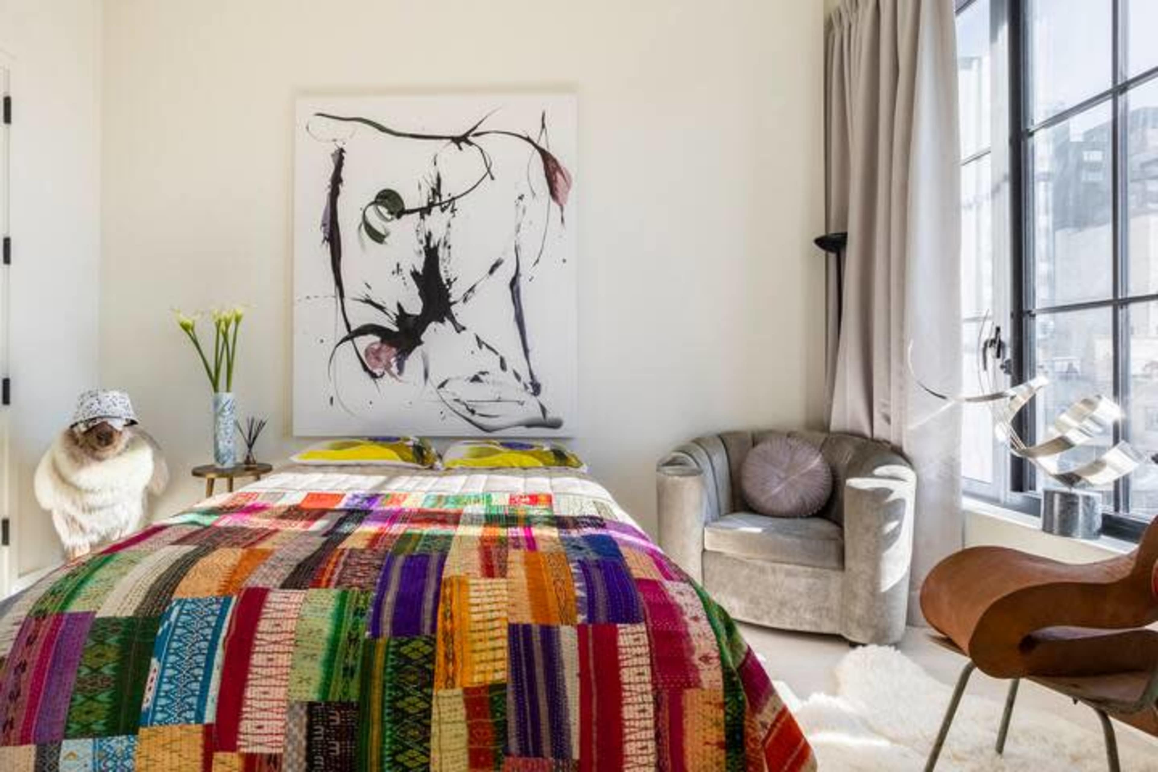 The image shows a modern bedroom featuring a colorful patterned bedspread, a contemporary armchair, and a large piece of artwork on the wall.