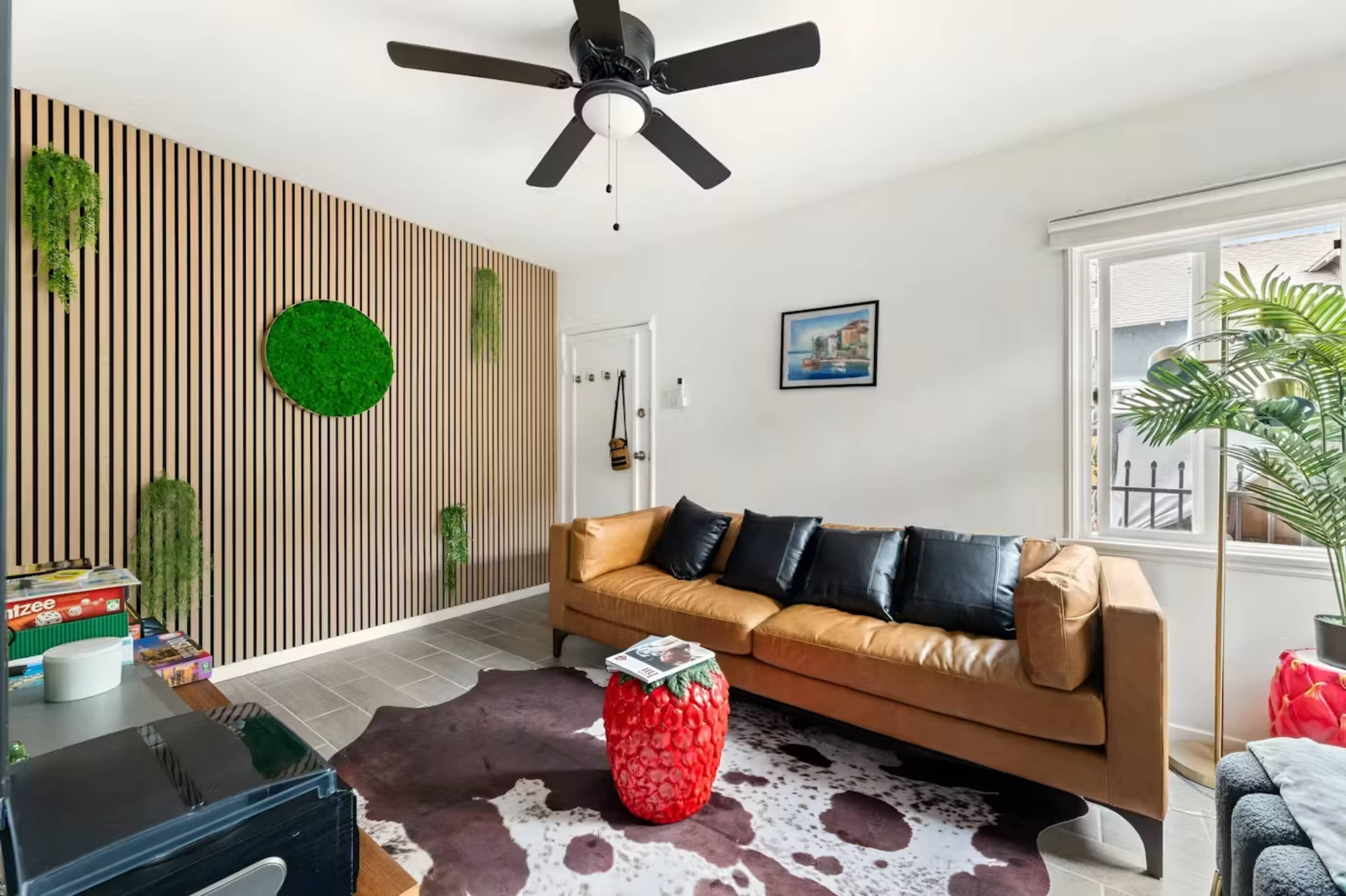 The living room features a brown sofa, a cowhide rug, a red strawberry-shaped table, and a green vertical garden on a wooden slat wall.