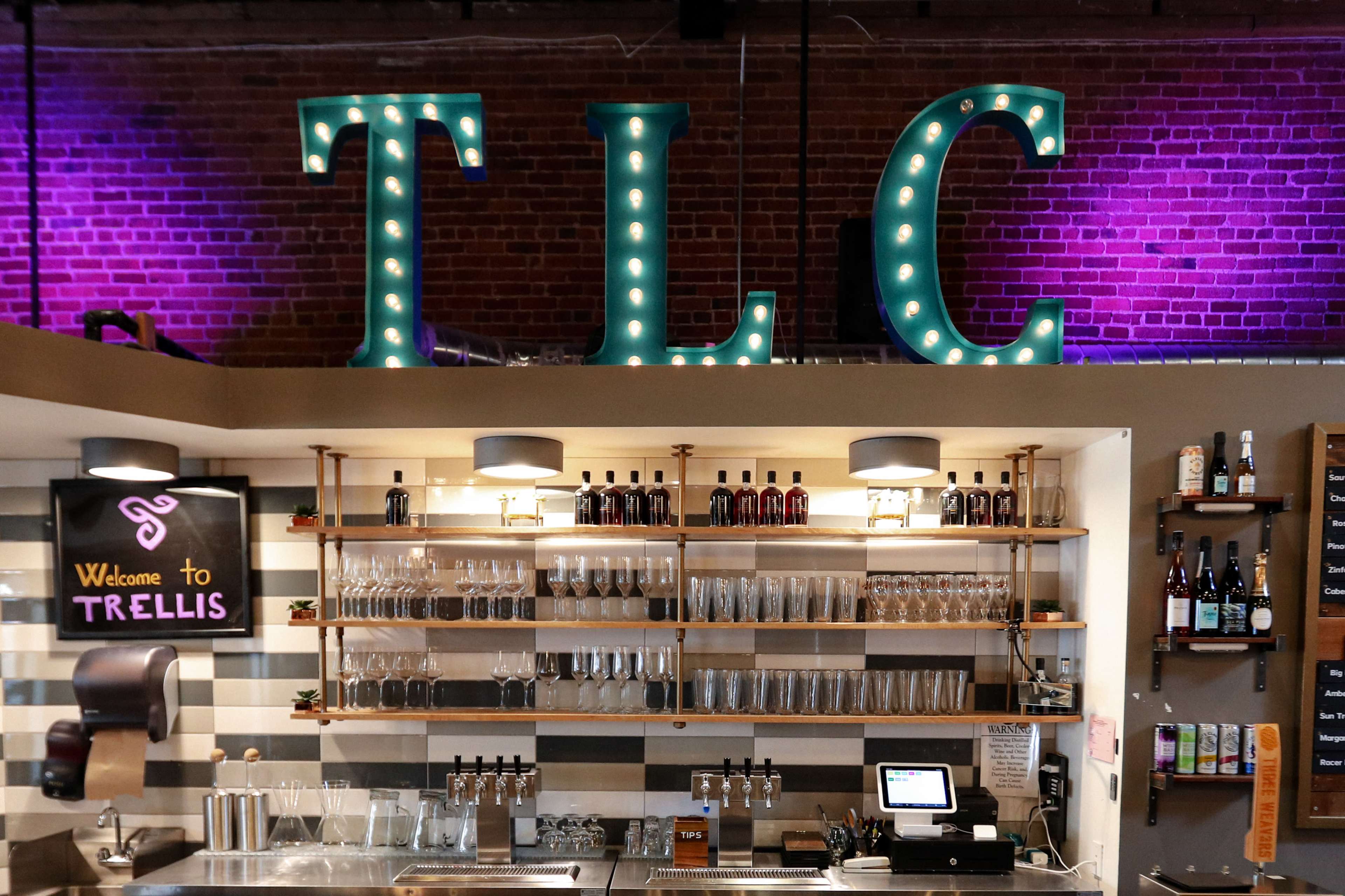 A bar area with a prominently lit "TLC" sign and shelves displaying bottles and glassware, along with a chalkboard that says "Welcome to Trellis."