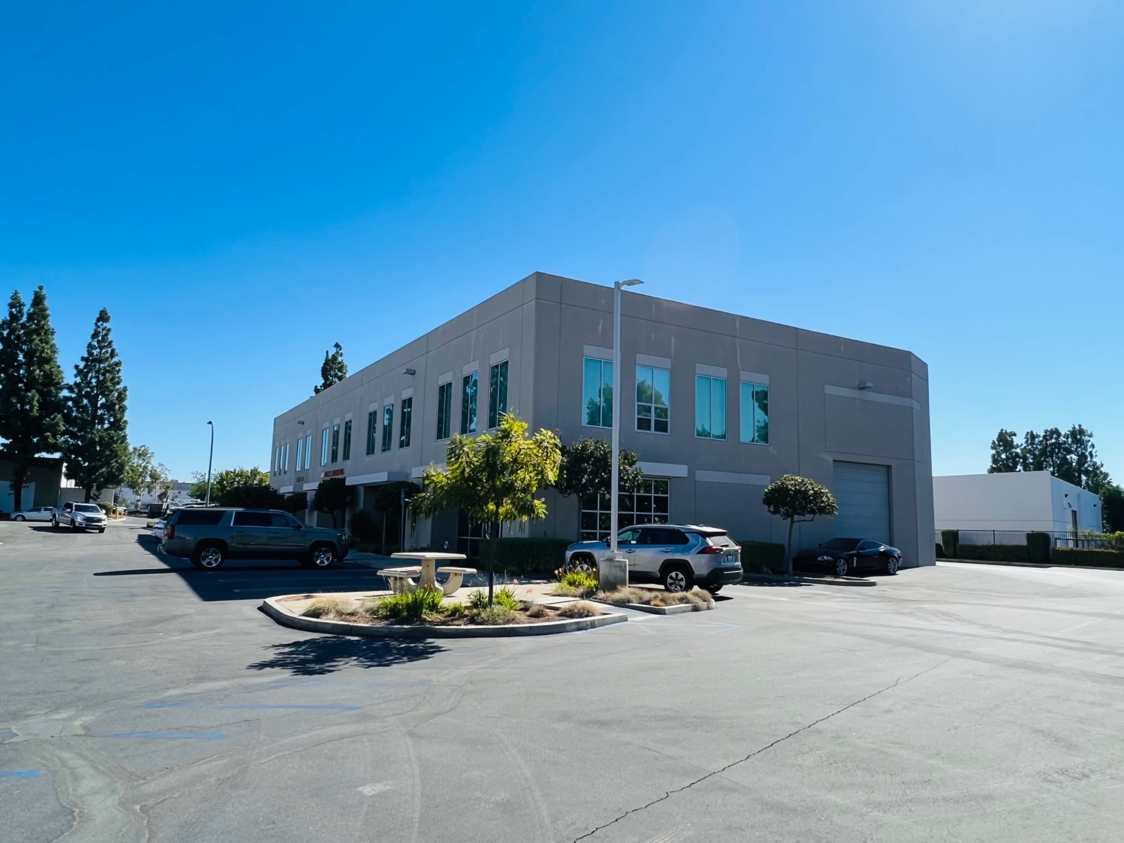 A two-story commercial building is situated in a parking lot with several cars and landscaping in front under a clear blue sky.