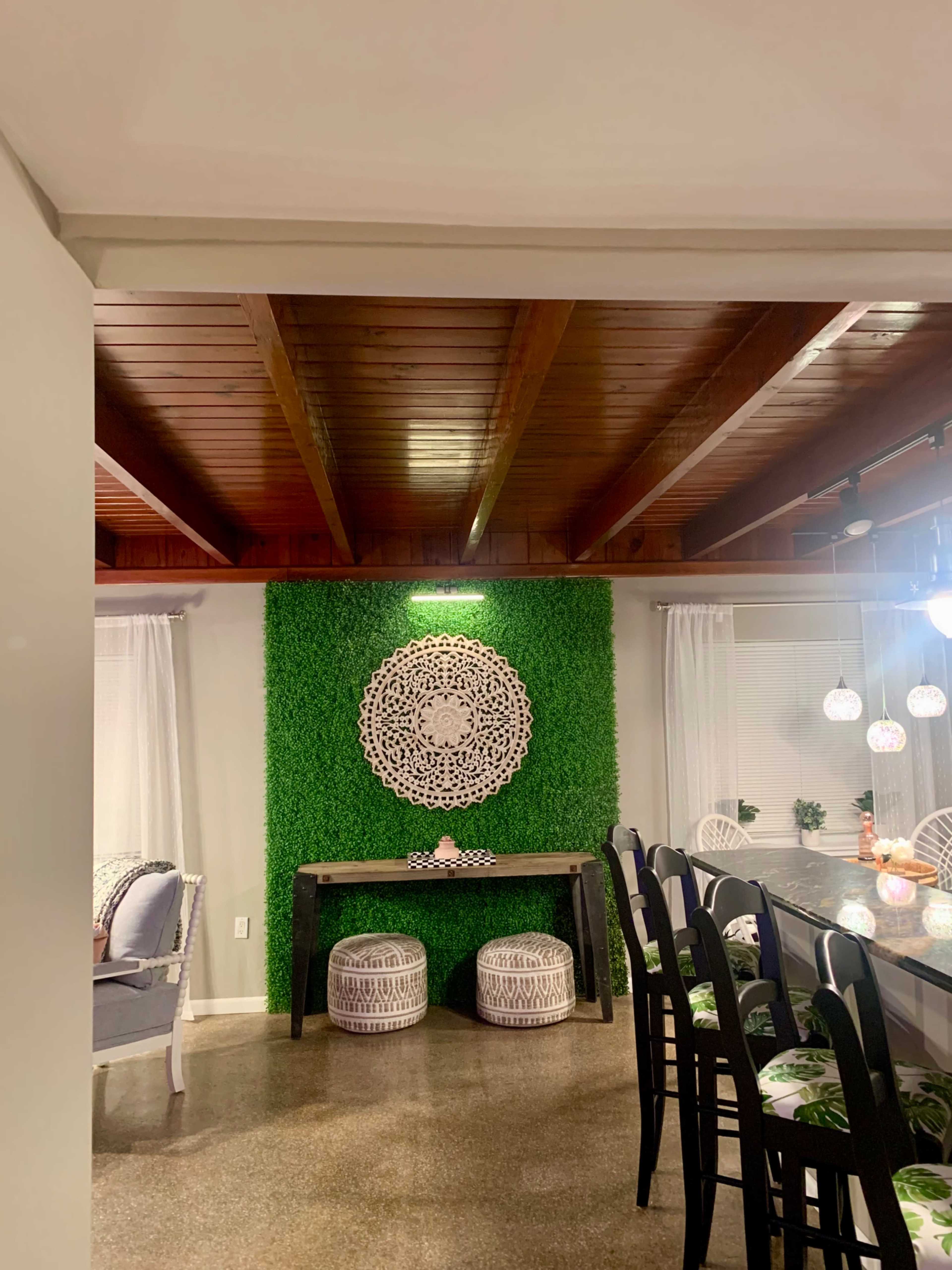 The image shows a well-lit dining area featuring a green textured wall with a decorative circular piece and a wooden table with two patterned stools.