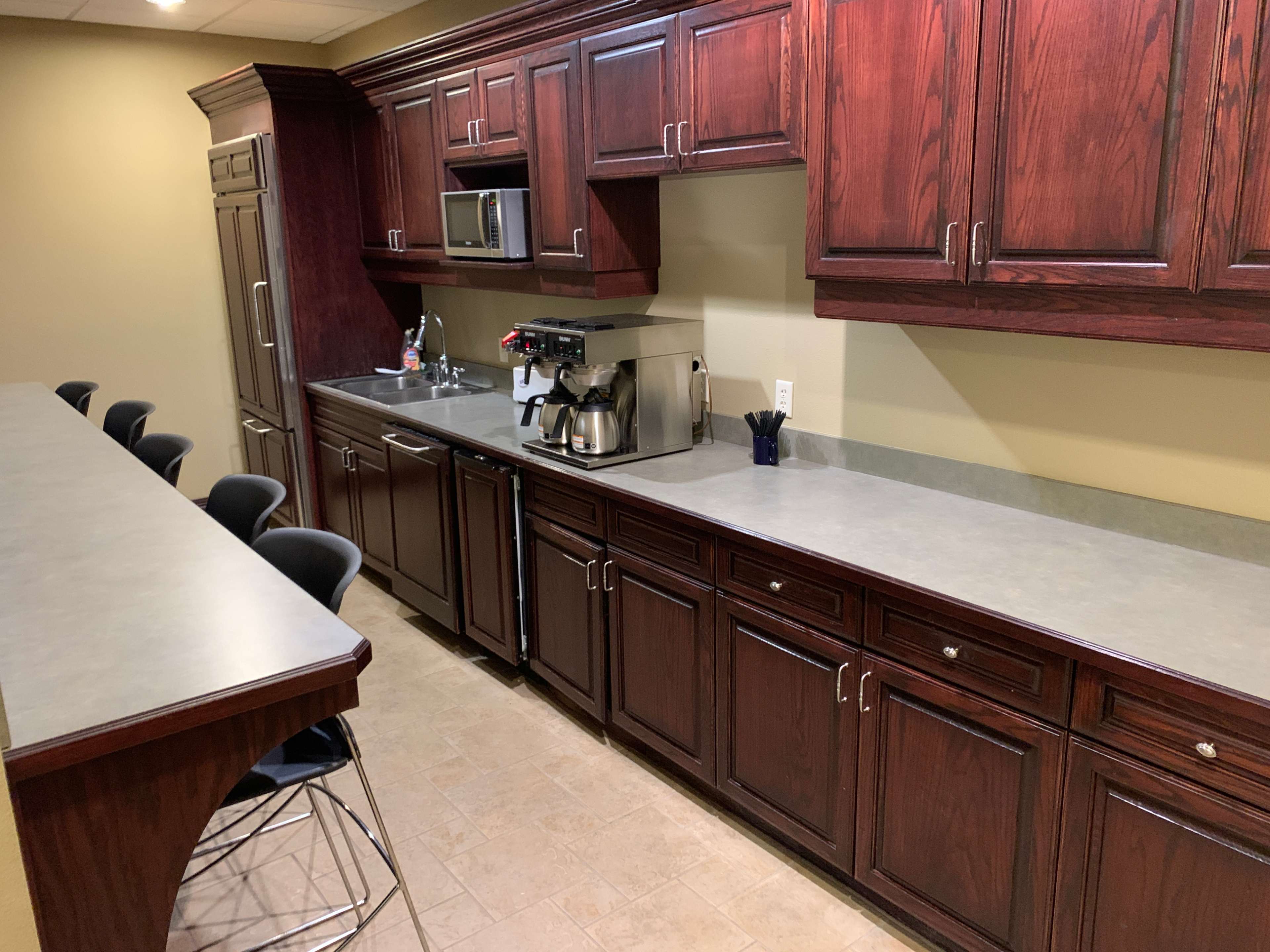 A modern kitchen with dark wooden cabinets, a countertop, and a coffee machine, positioned next to a bar with high stools.