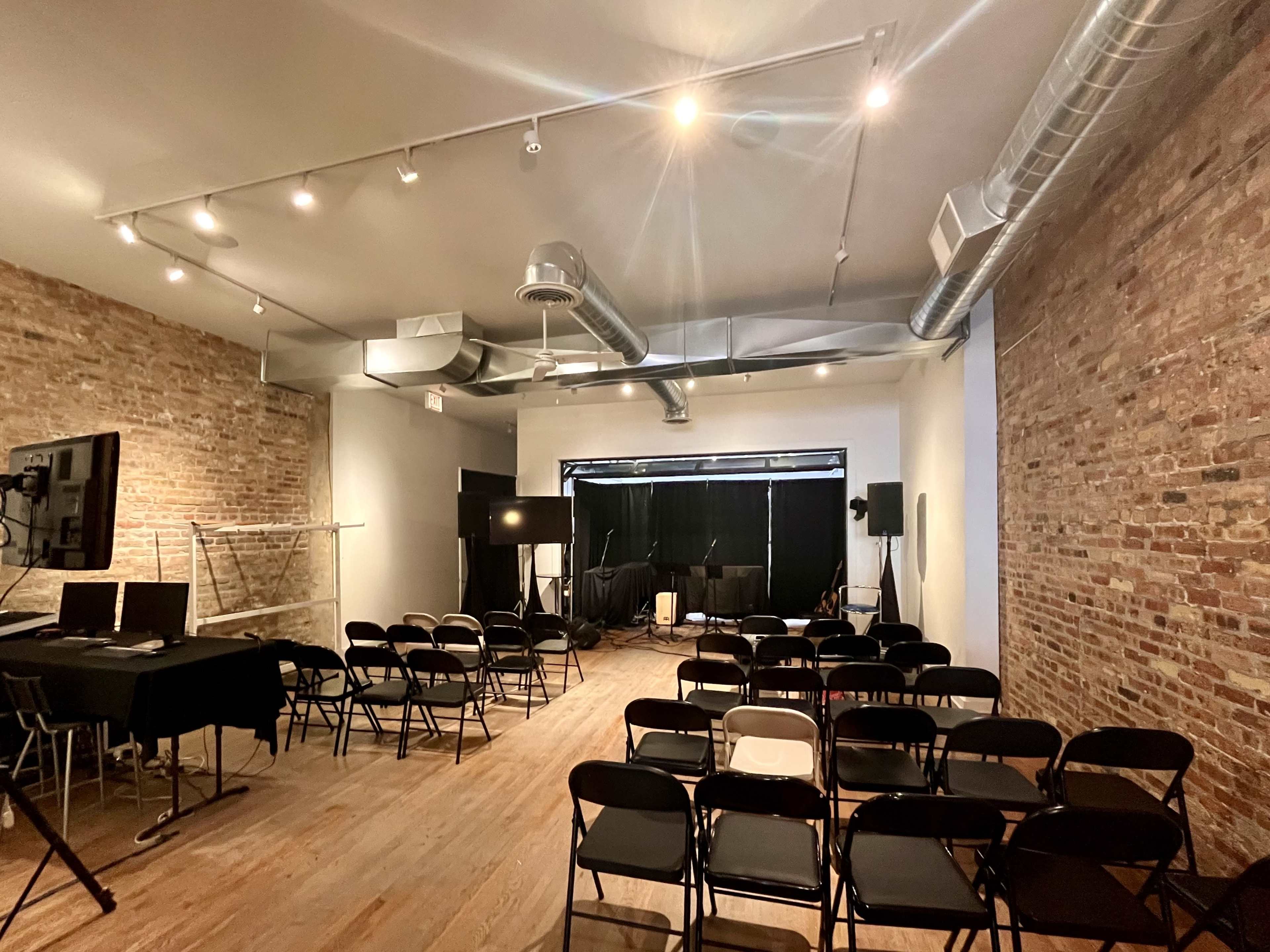 A minimally furnished event space with wooden flooring, exposed brick walls, and rows of black chairs set up for an audience facing a stage area.