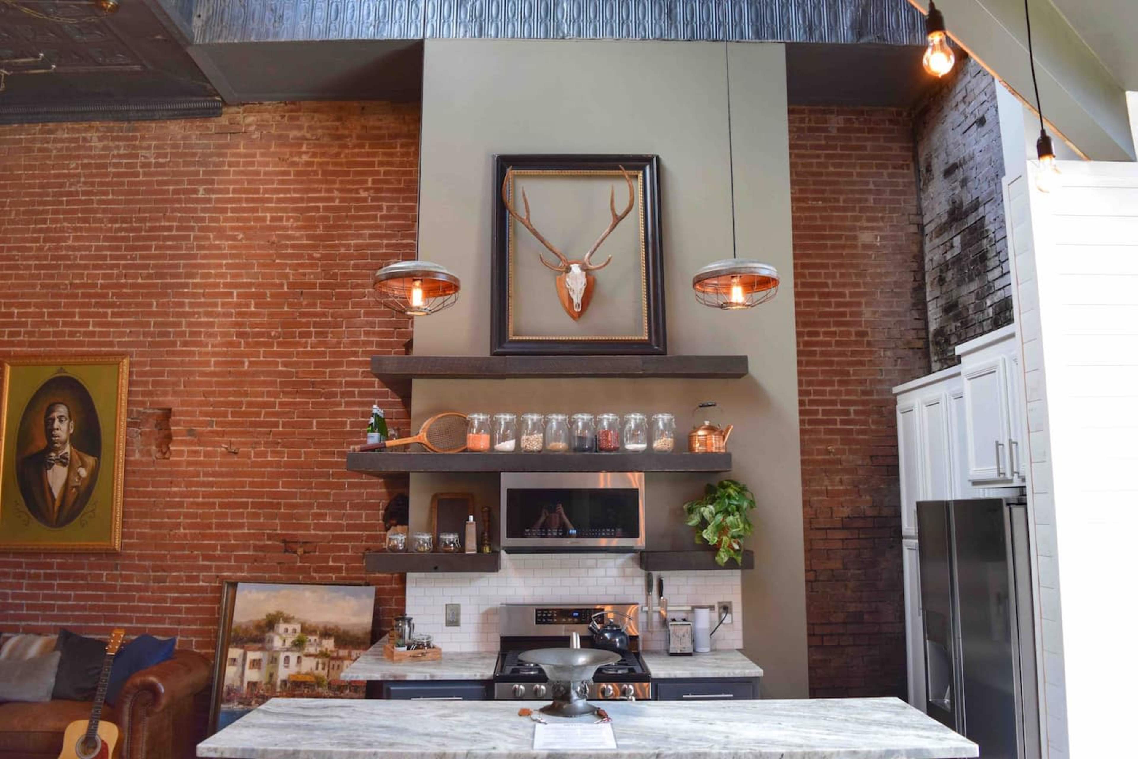A modern kitchen with exposed brick walls, a central island, and a decorative mounted deer head above the shelves.